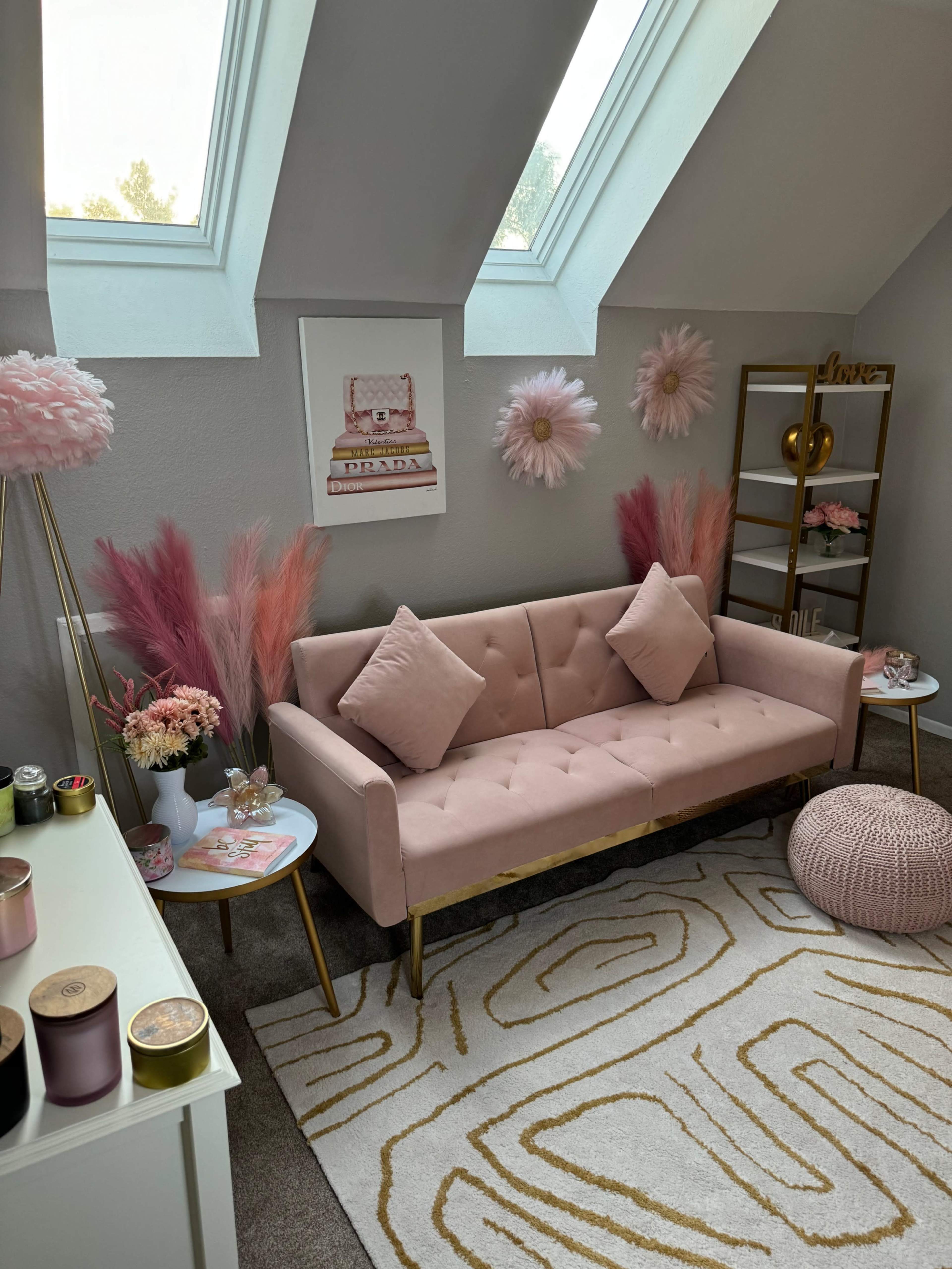 The image shows a stylish room featuring a pink sofa with golden legs, adorned with decorative pillows, surrounded by various decorative elements like a bookshelf, wall art, and soft accent pieces.