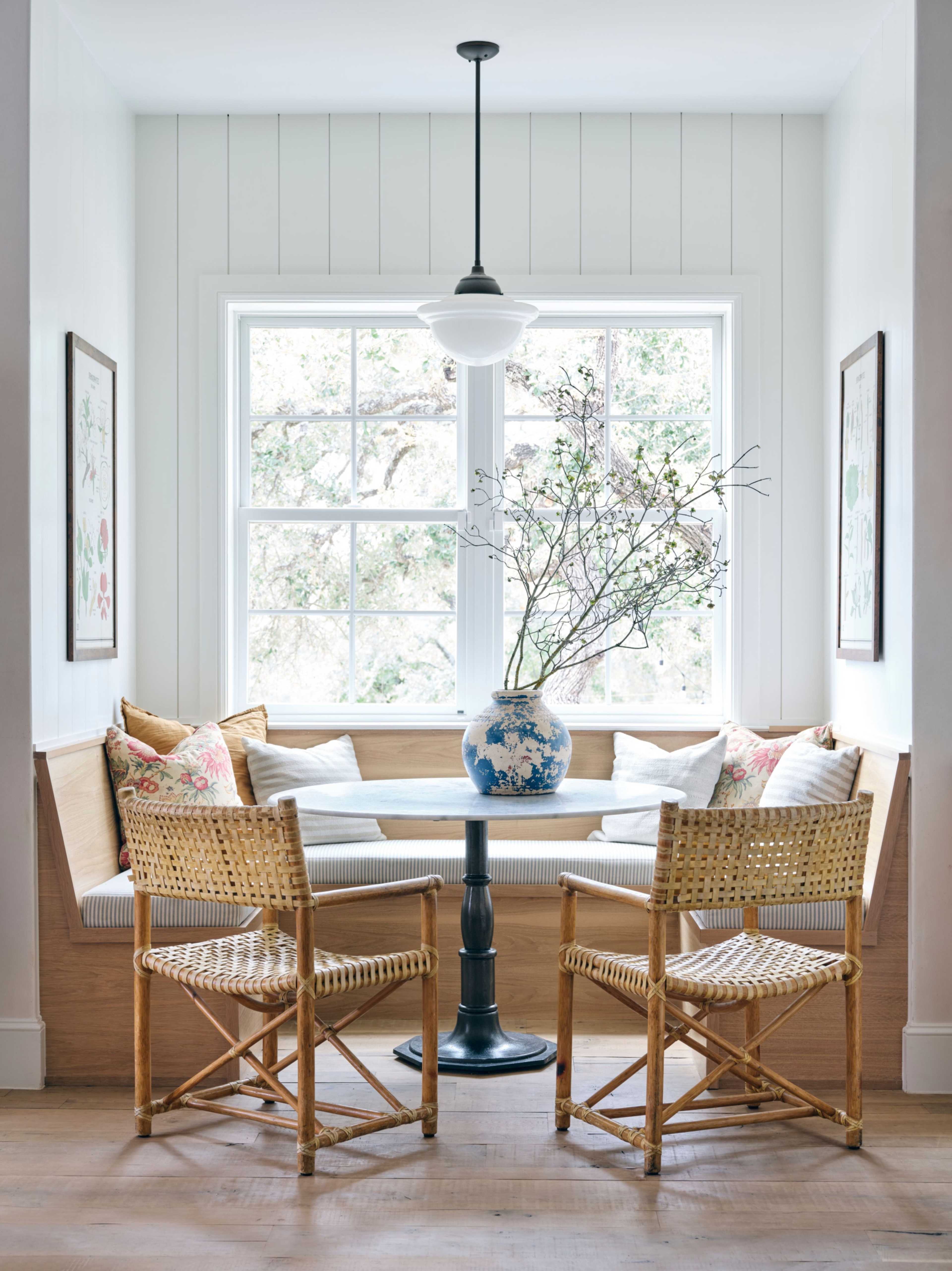 A cozy nook features a wooden bench with cushions, a round table, and two rattan chairs under a pendant light, beside a window displaying a decorative vase with branches.