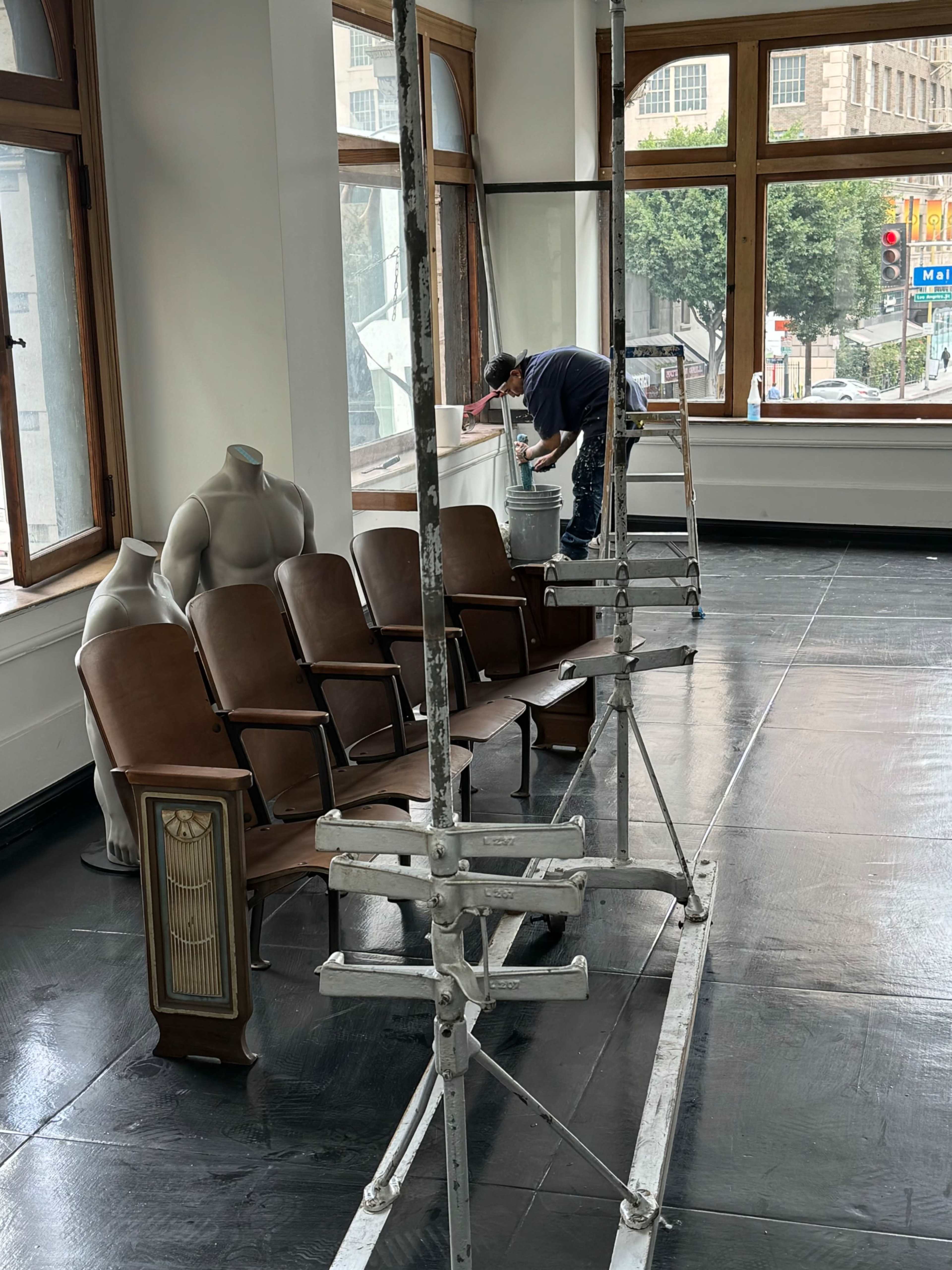 A worker on a ladder paints a wall in a room with vintage-style seating and scaffolding in the foreground.