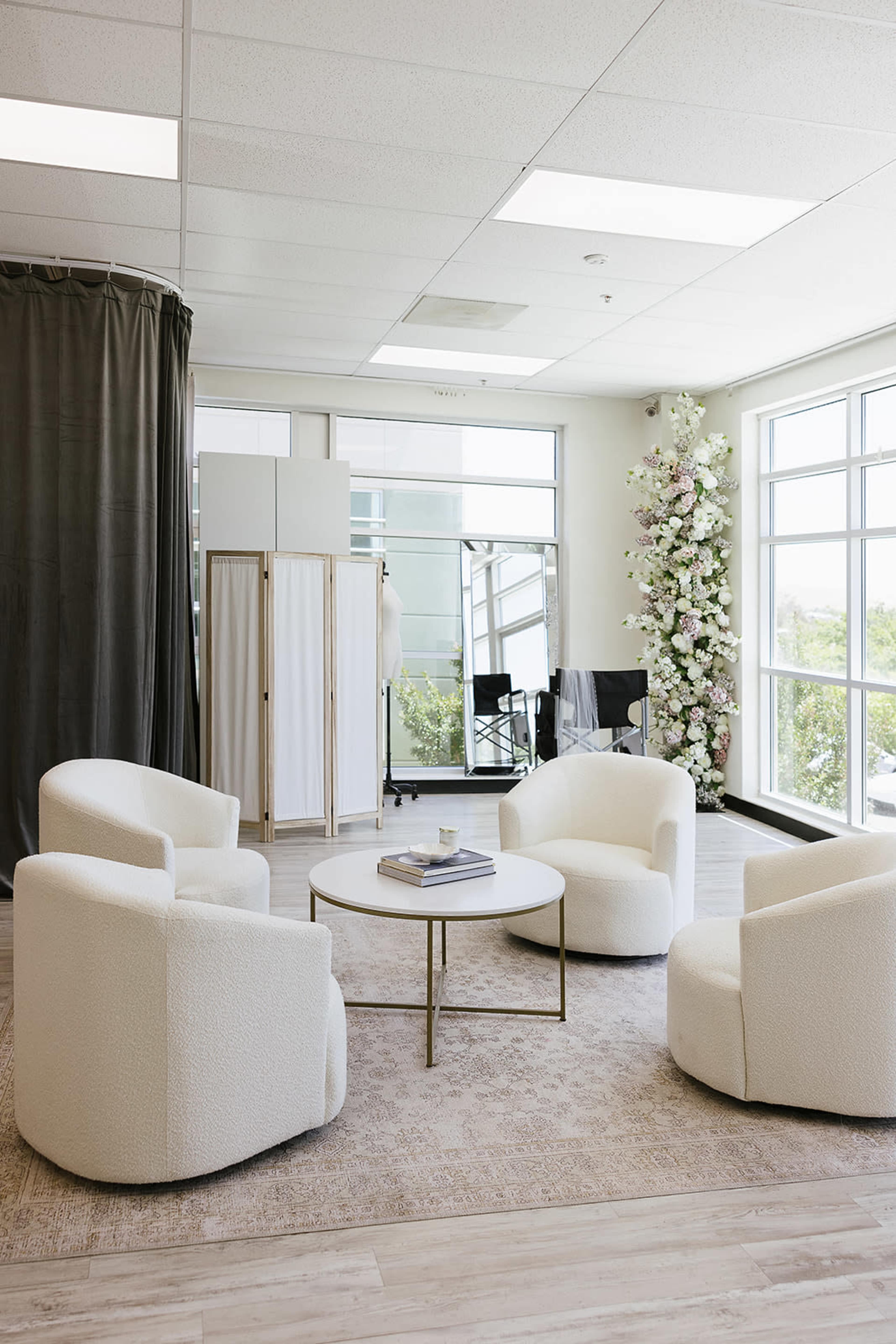 The image shows a bright, modern lounge area with four white, upholstered chairs arranged around a circular coffee table, large windows, and decorative plants.