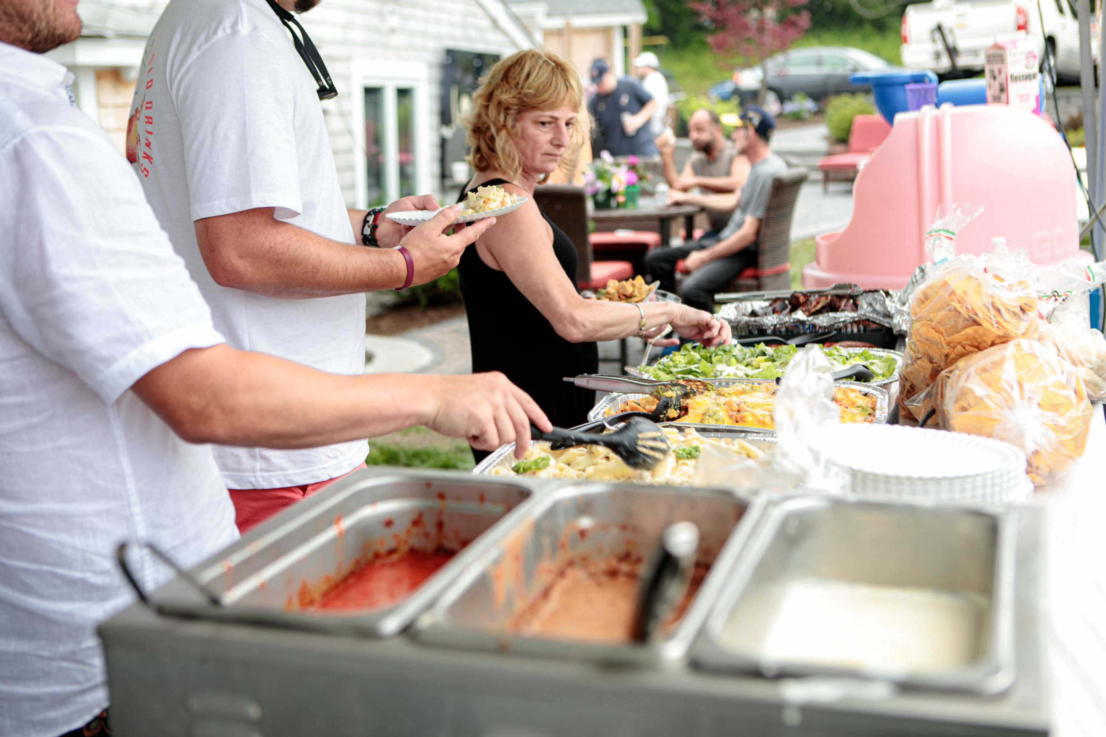 A buffet table is set up outdoors, with people serving themselves food as others gather nearby to enjoy the meal.