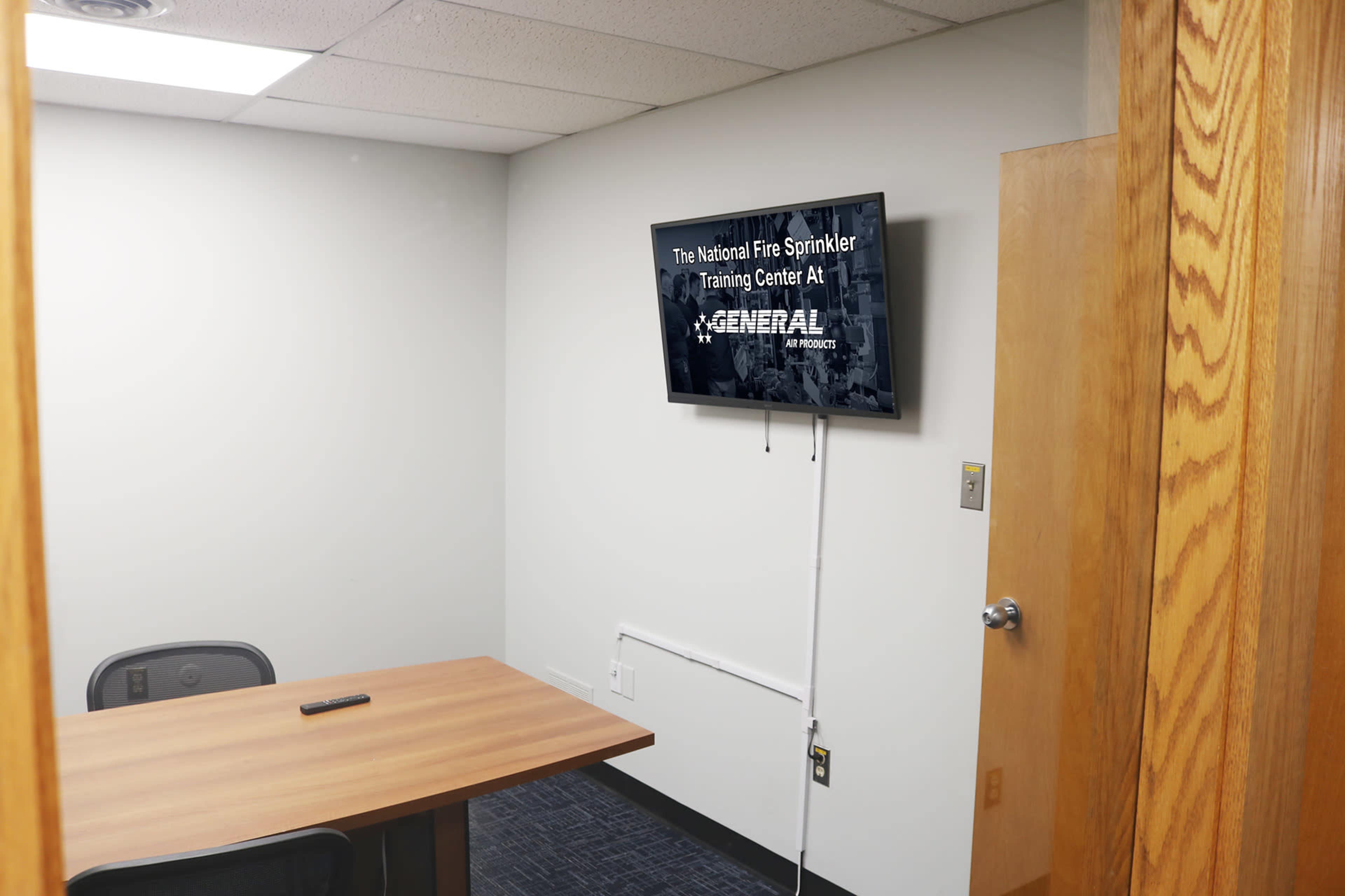 The image shows a training room with a wooden table, a chair, and a wall-mounted television displaying the title "The National Fire Sprinkler Training Center."