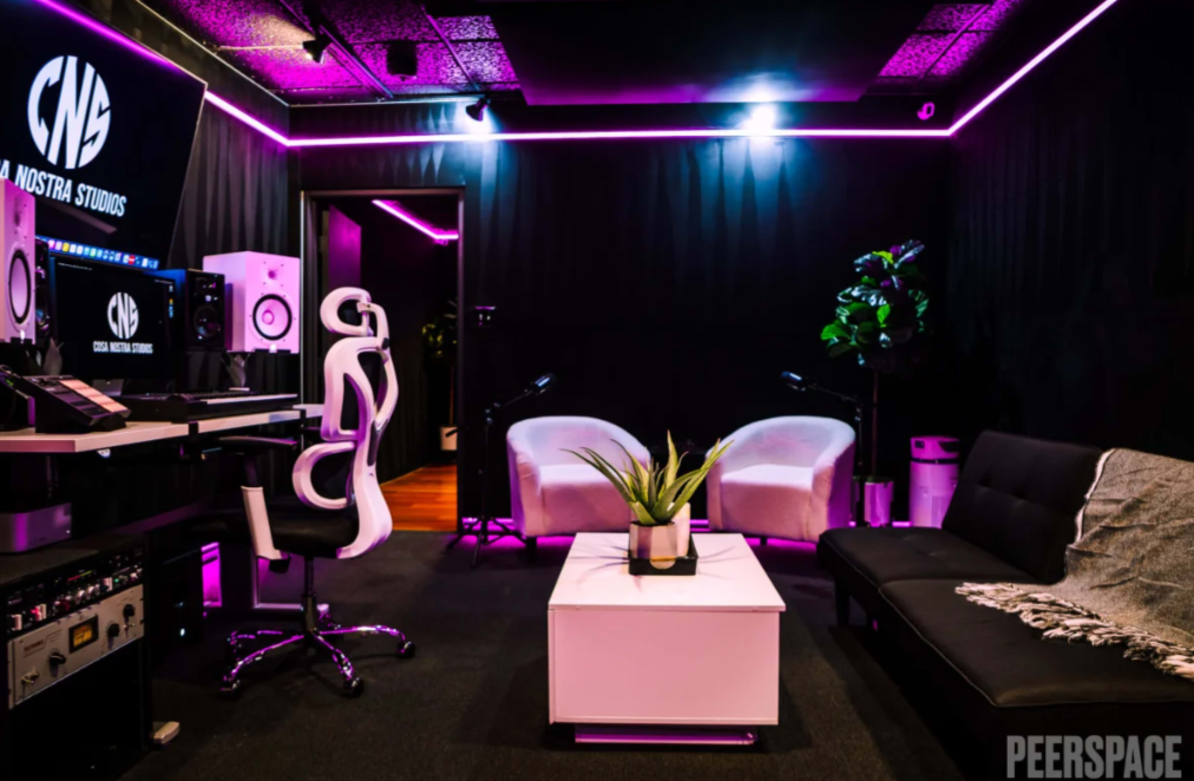 The image shows a modern recording studio with a black and pink color scheme, featuring a workstation, seating area, and decorative plants.