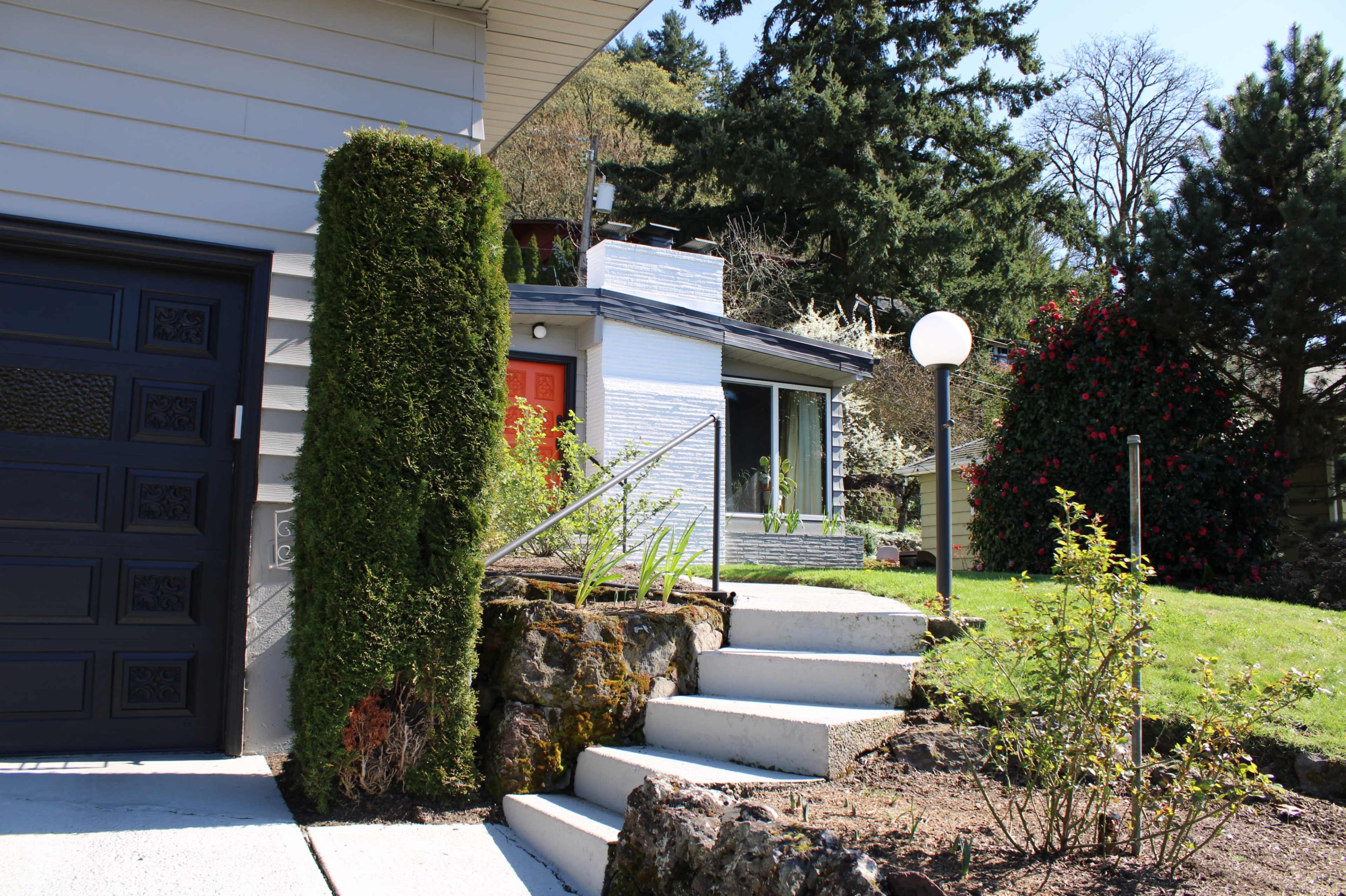 The image shows a well-maintained house with a neatly landscaped yard, featuring a set of white steps leading to a front door surrounded by greenery.