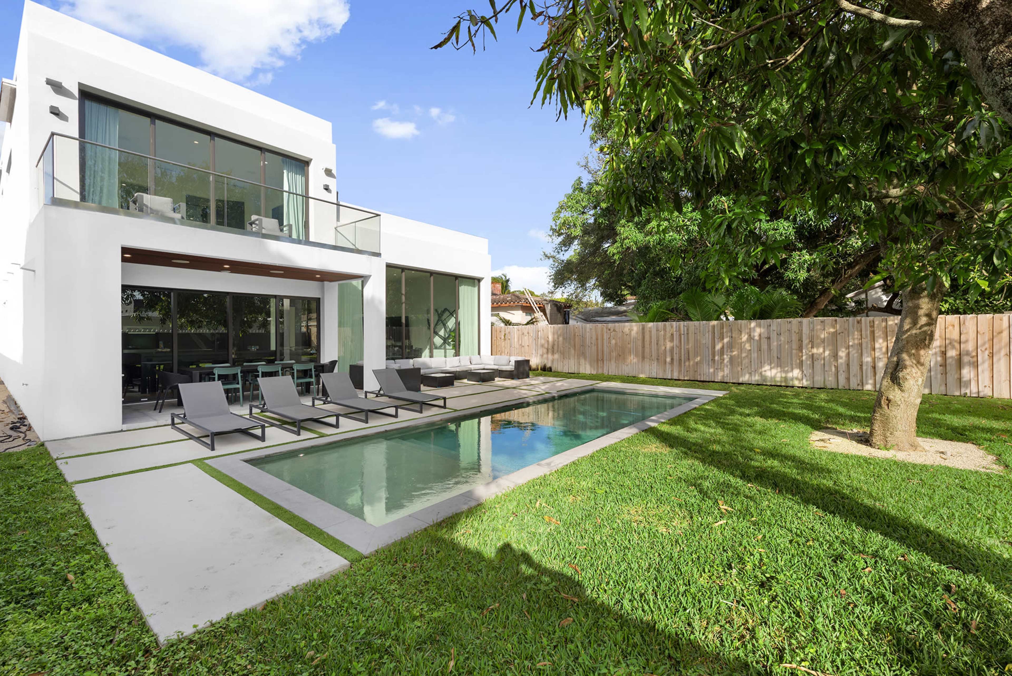 The image shows a modern two-story house with large windows, a swimming pool, and a landscaped lawn surrounded by a wooden fence.