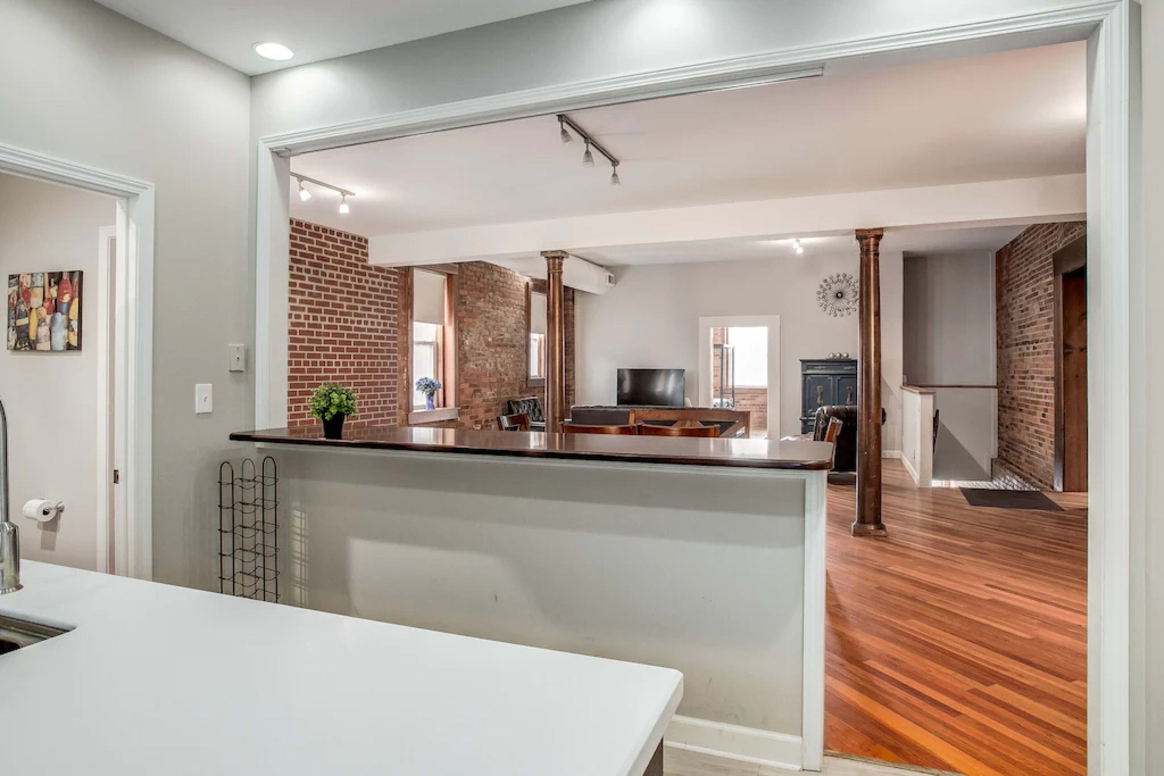 A view of an open-concept living space with a kitchen island, hardwood flooring, exposed brick walls, and a seating area visible through an archway.