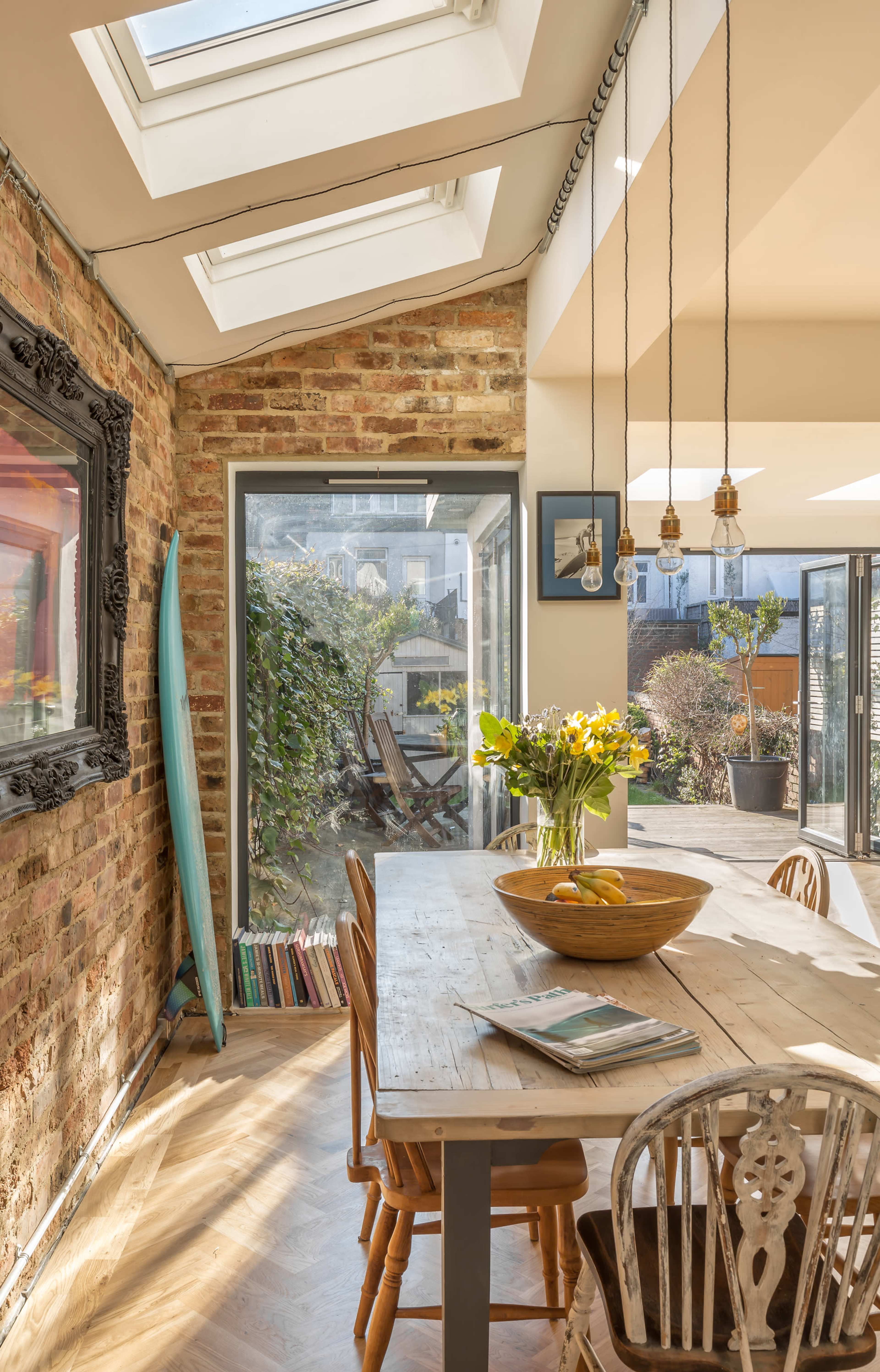 A wooden dining table sits in a sunlit kitchen with large skylights and a brick wall, while a vase of flowers and a bowl of fruit decorate the table.