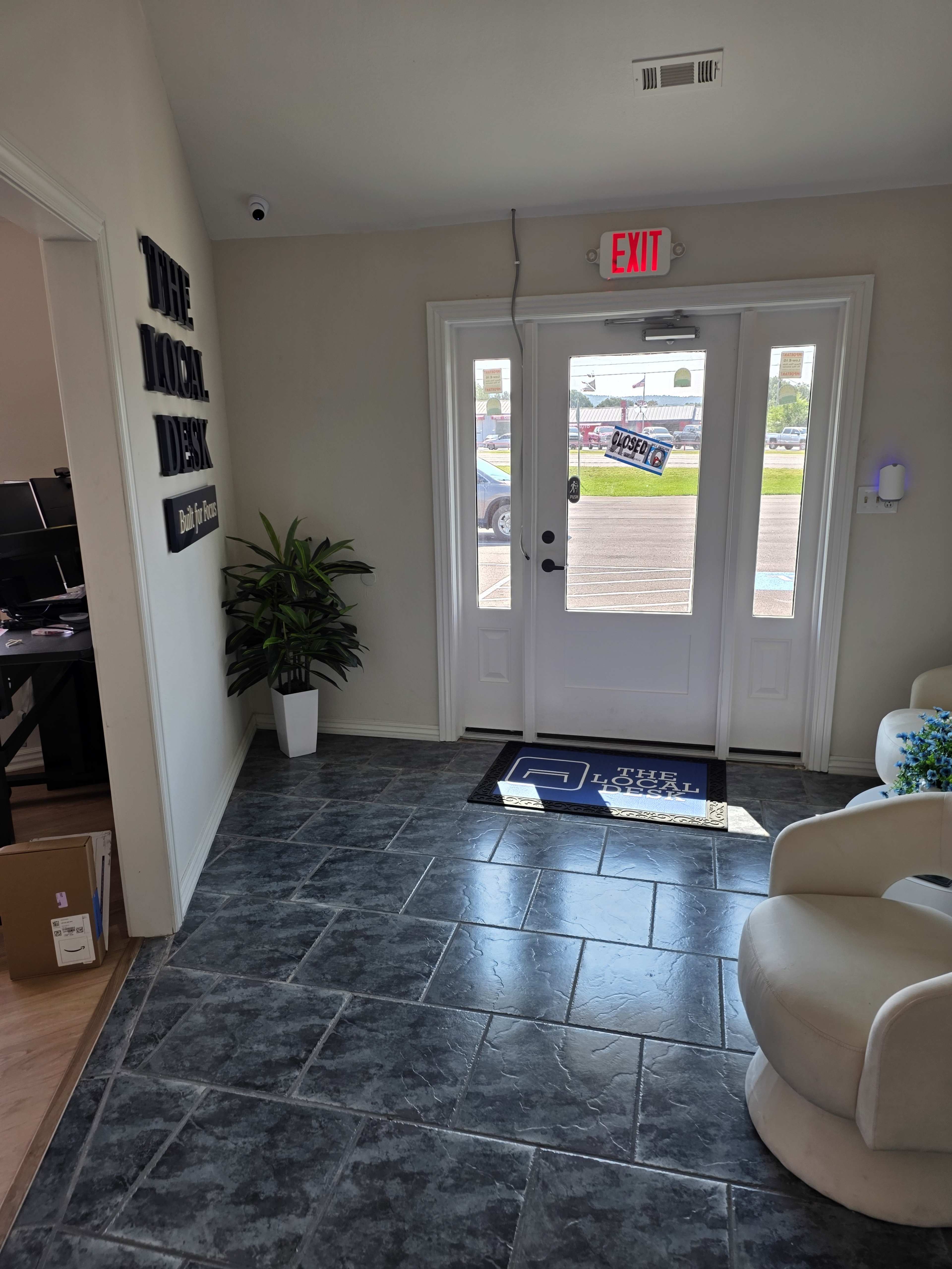 The image shows a brightly lit reception area featuring slate flooring, a potted plant, and a door leading outside, with a sign labeled "THE LOCAL DESK" on the wall.