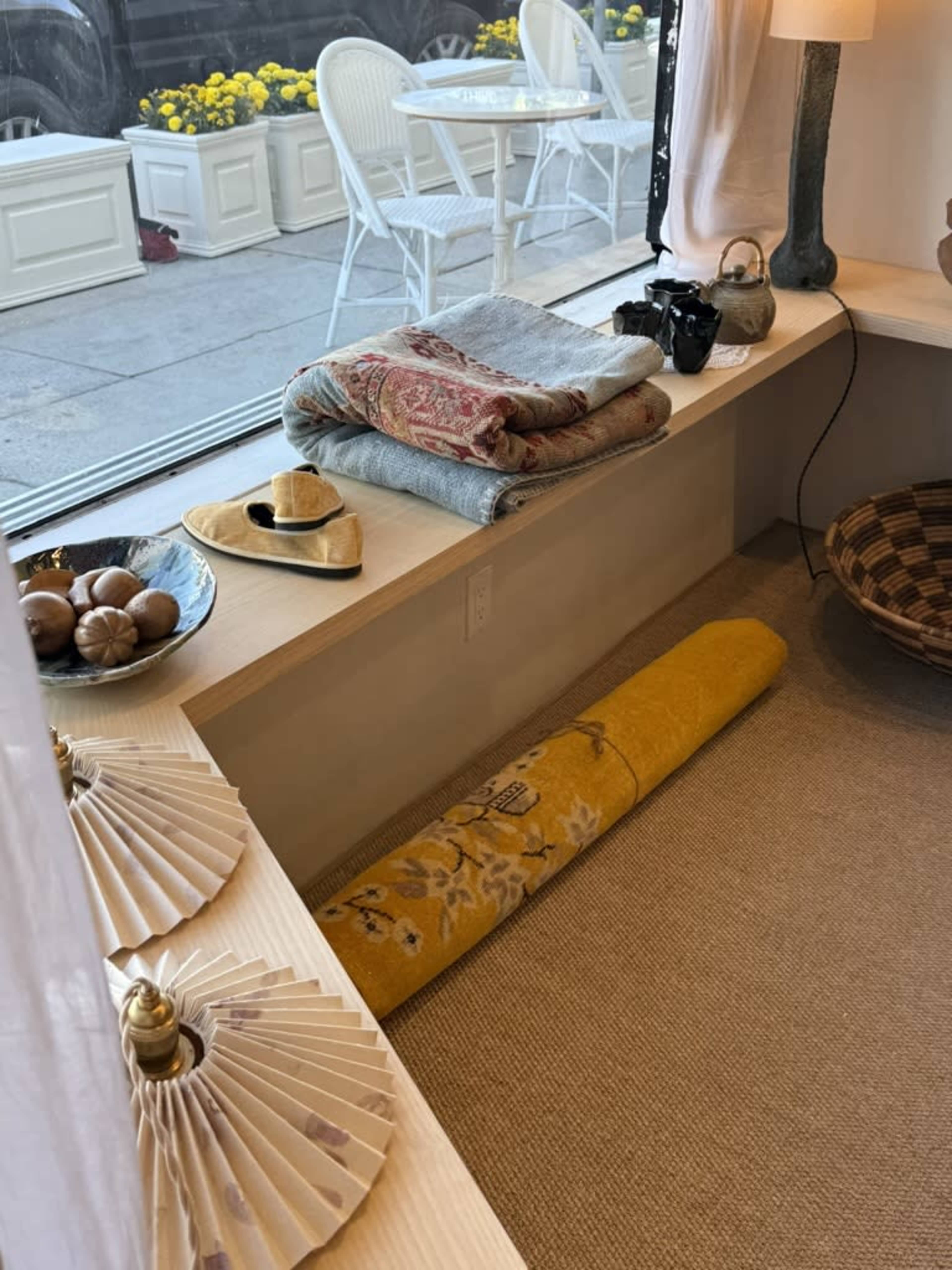 A windowsill features neatly arranged textiles, a camera, a straw hat, and decorative items, with yellow flower planters visible outside.