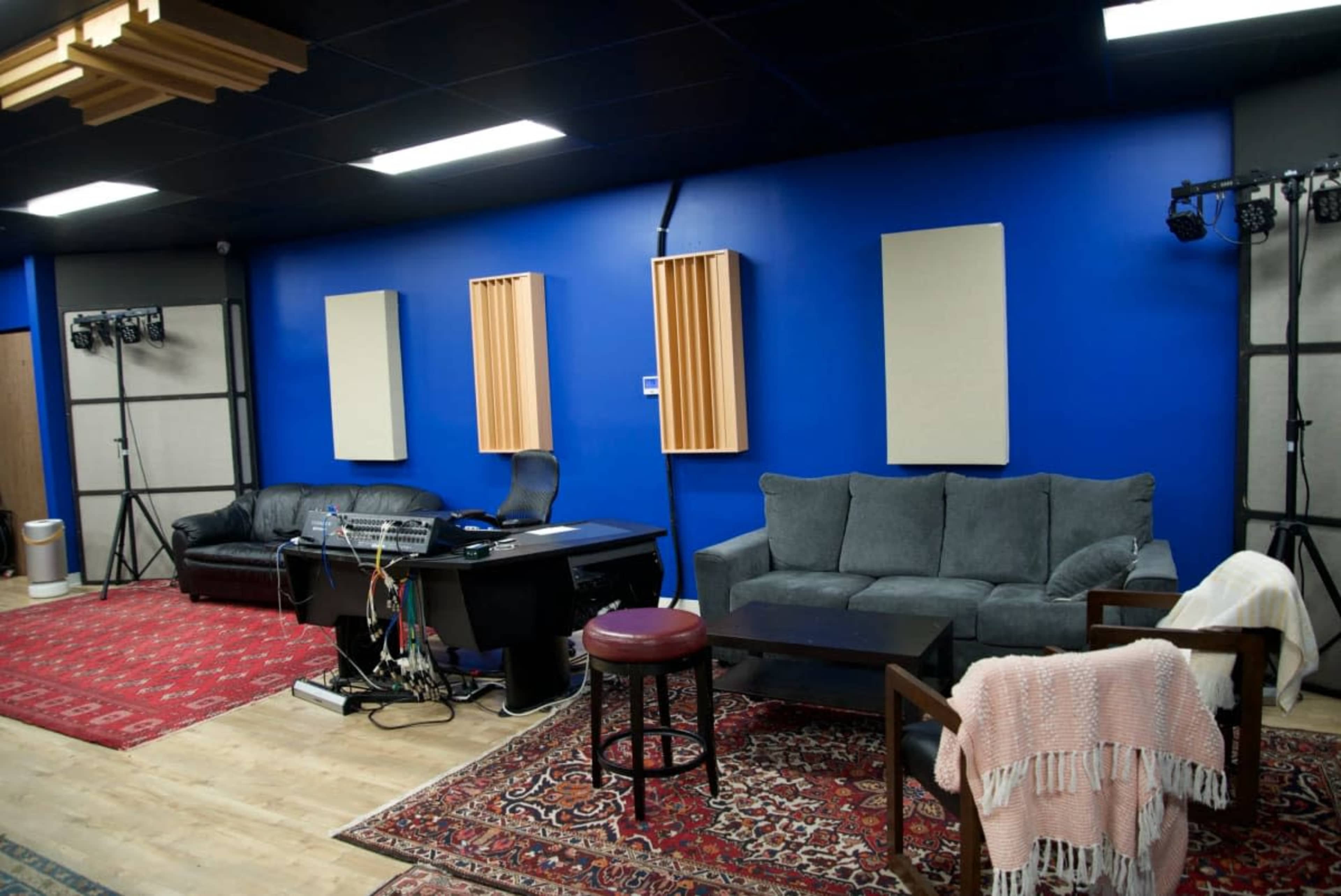 The image shows a recording studio with blue walls, soundproof panels, a mixing desk with cables, two sofas, and a small table, set against a wooden floor with area rugs.