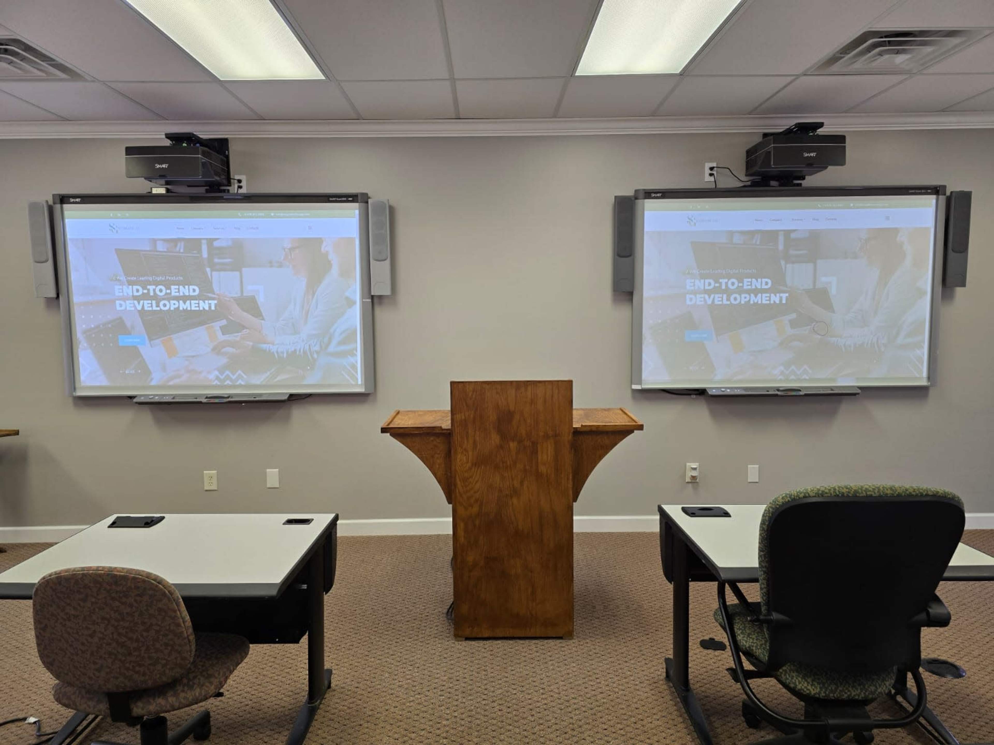 A conference room with two large screens displaying "END-TO-END DEVELOPMENT," a wooden podium at the front, and tables arranged for seating.