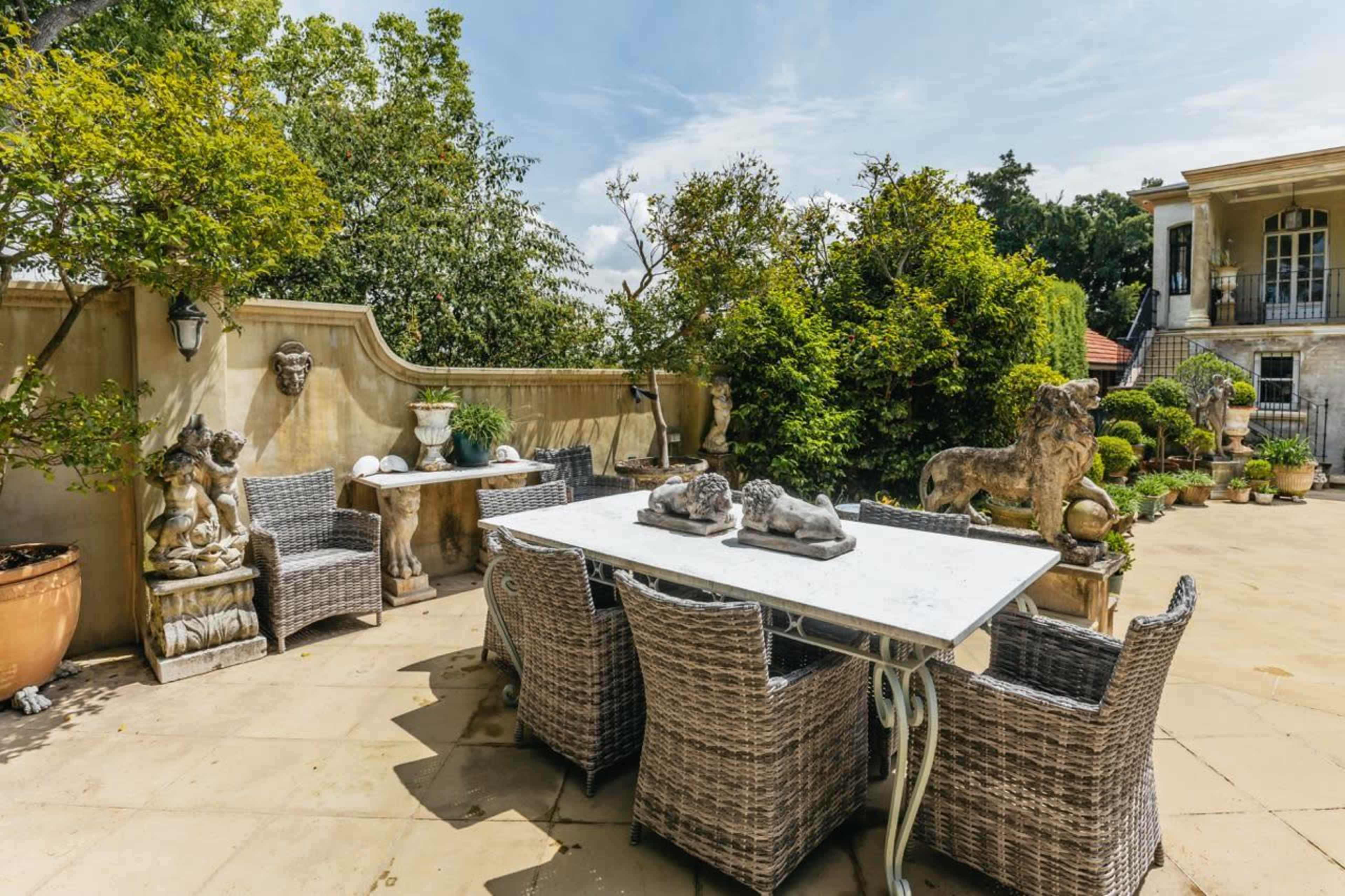 A spacious patio with a white table and wicker chairs surrounded by decorative plants and stone sculptures.