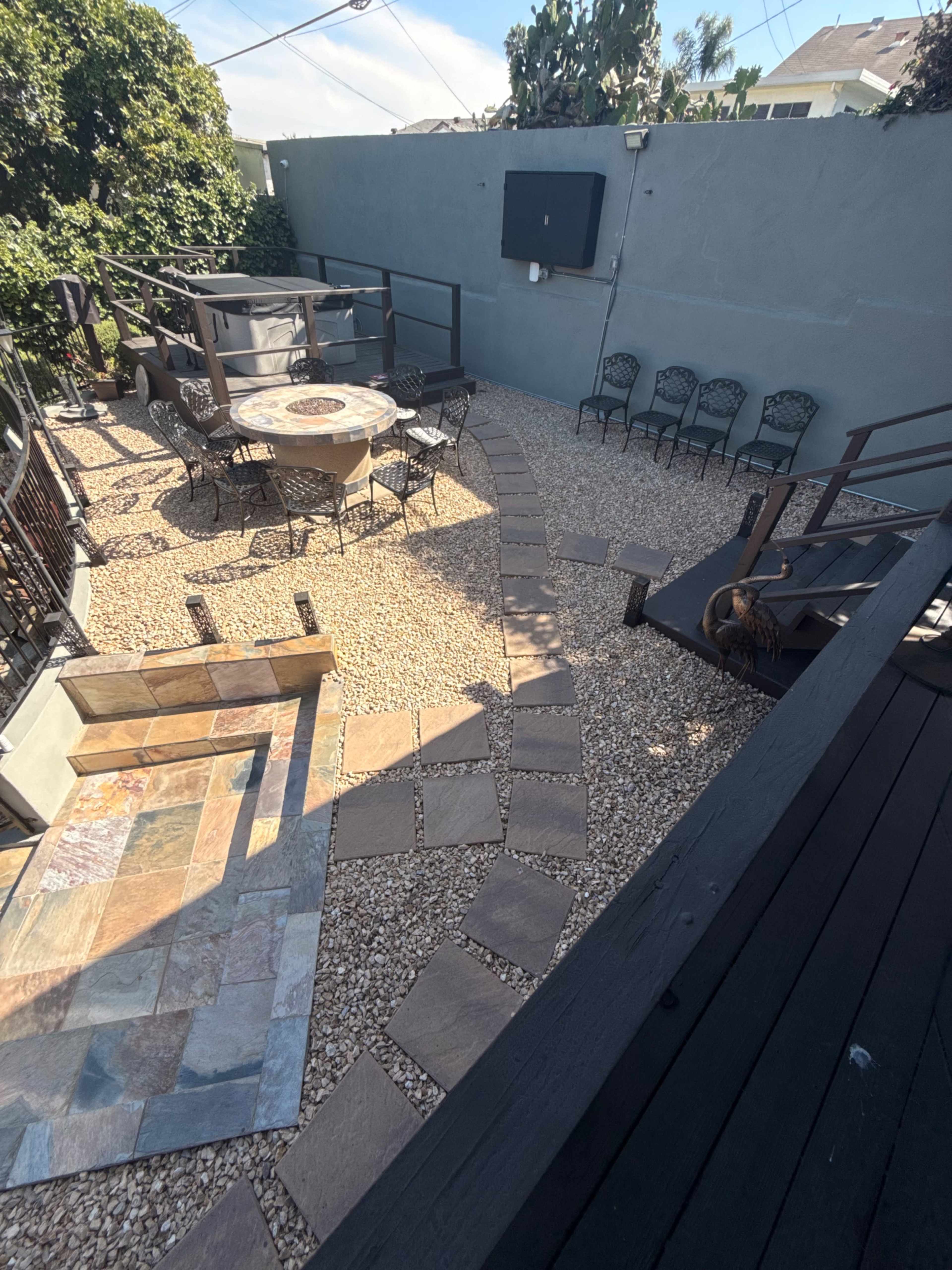The image shows a landscaped outdoor area with a stone path, a circular seating arrangement around a fire pit, and a television mounted on the wall.