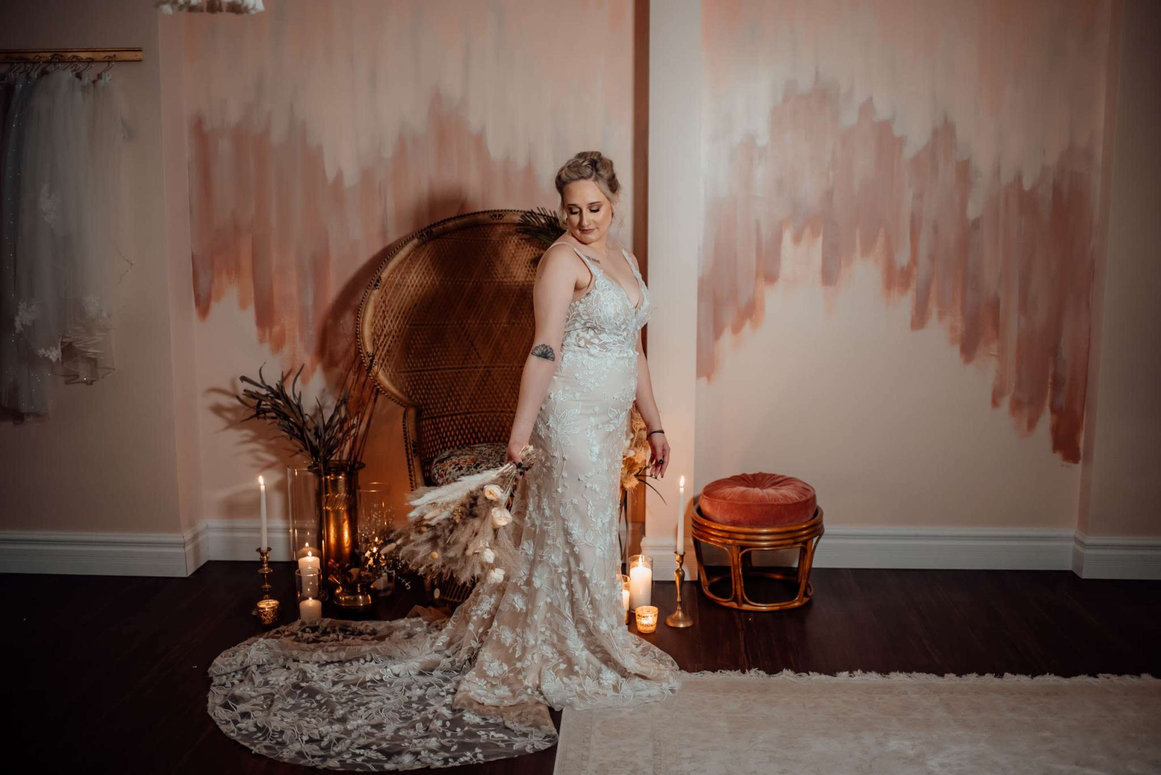 A woman in a detailed wedding gown poses with a bouquet in a softly lit room decorated with candles and a bohemian-style chair against a pastel wall.