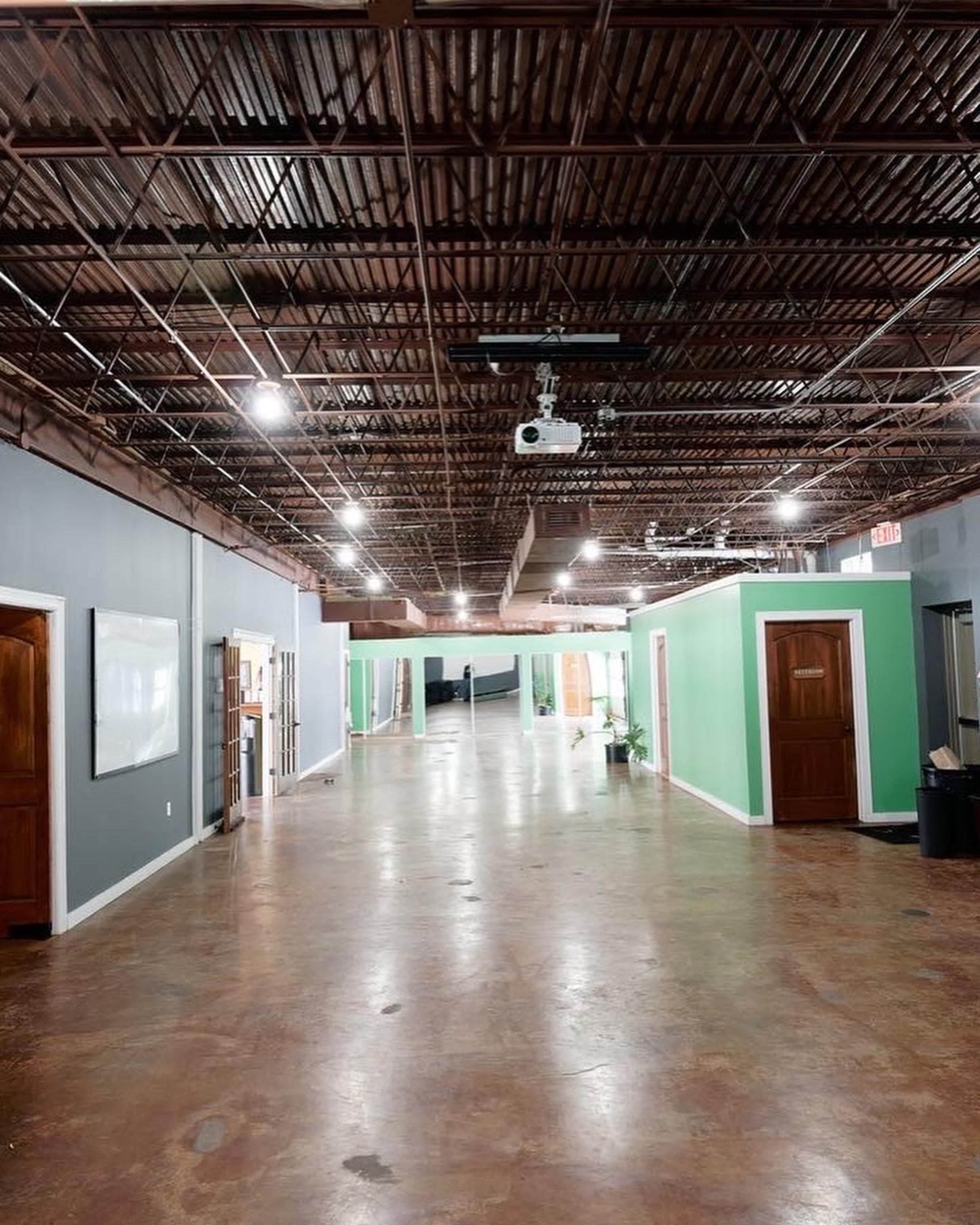 The image shows a spacious hallway with green and gray walls, featuring several doors and polished concrete flooring under a metal ceiling.