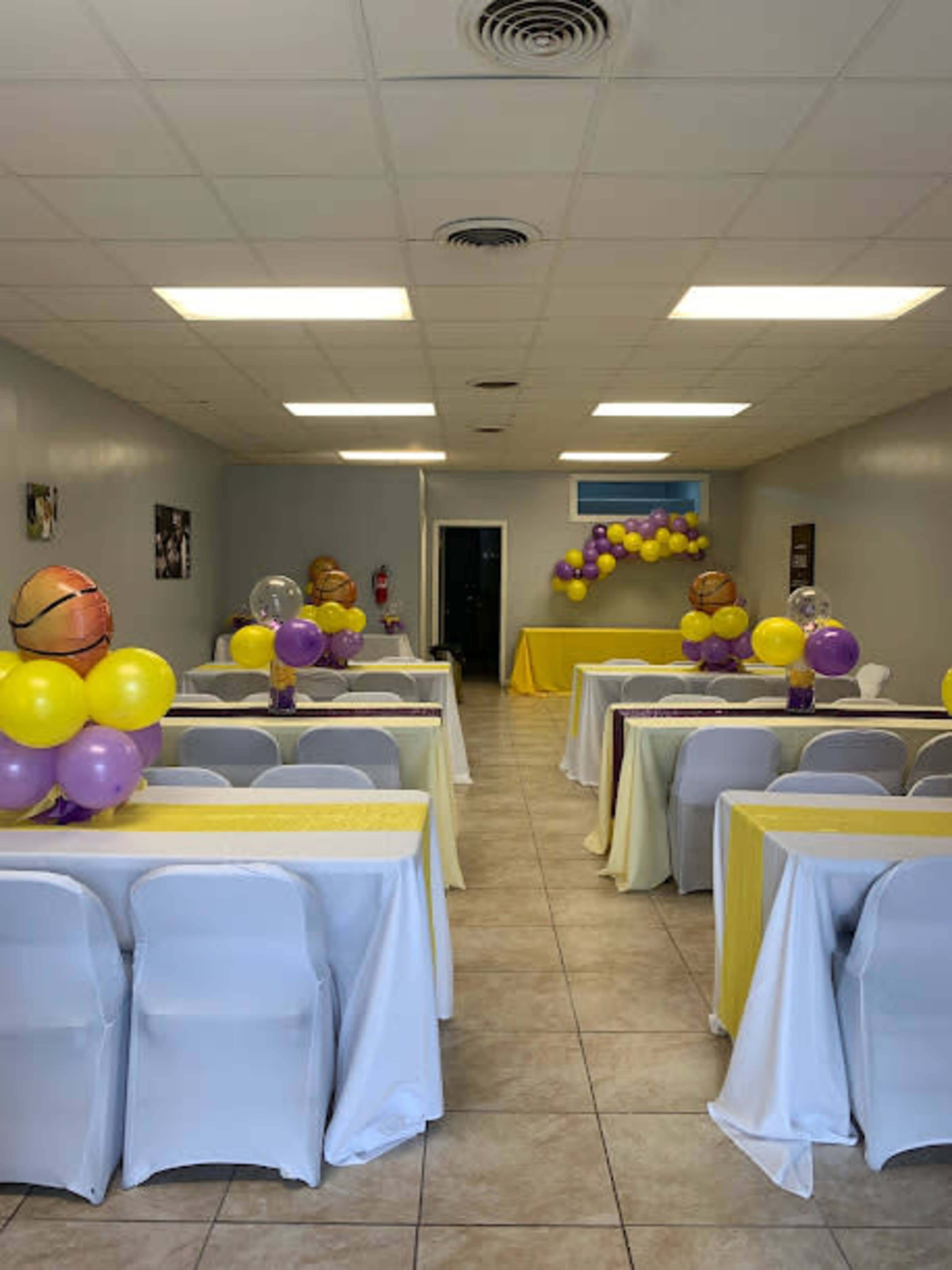 A banquet hall arranged with tables covered in yellow and purple tablecloths, decorated with balloons, and a small display table at the back.