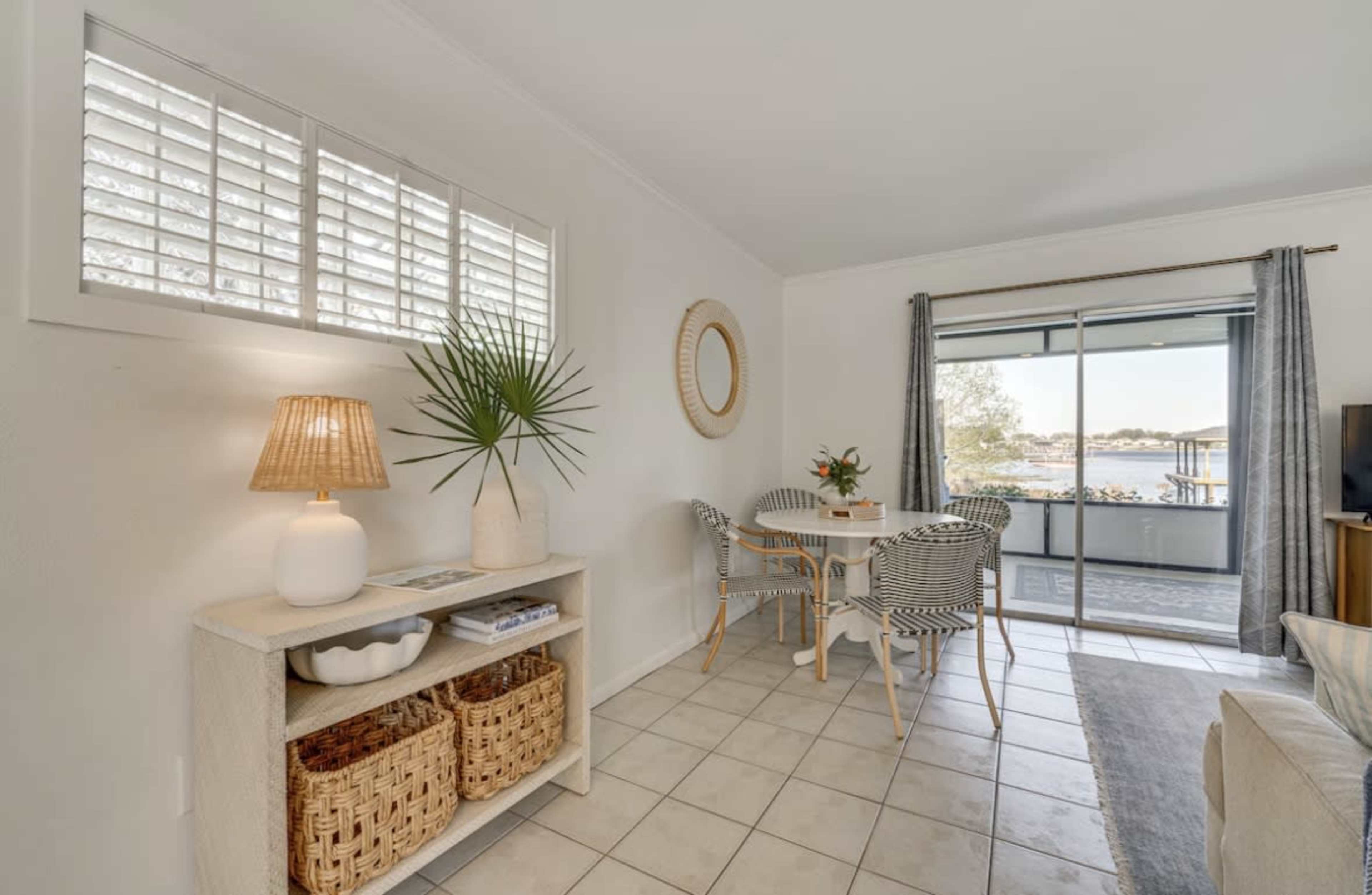 The image shows a bright living space featuring a dining area with two chairs and a round table, a console table with a lamp and decorative plant, and large windows overlooking a waterfront view.