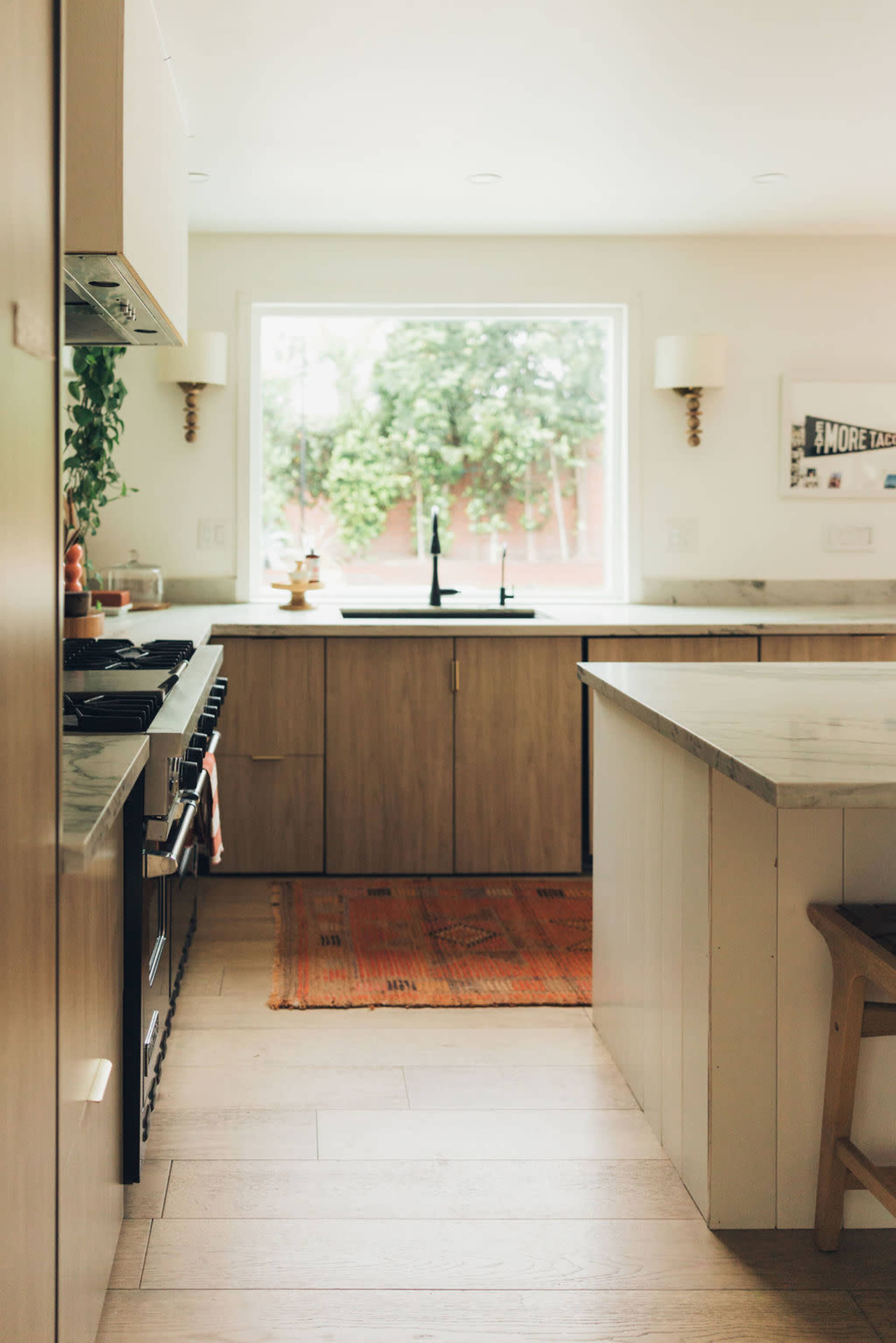 A bright kitchen features wooden cabinetry, a large window with greenery outside, and a patterned rug on the floor.
