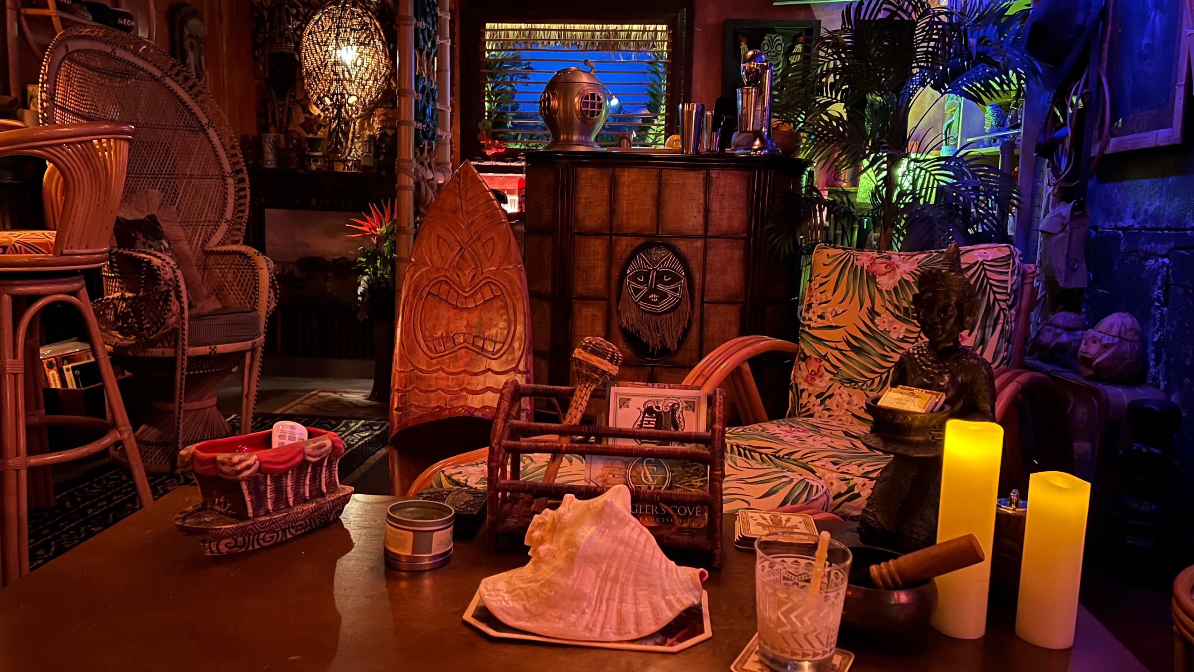 The image shows a dimly lit room decorated with tropical-themed furniture, a carved wooden figure, and various objects arranged on a wooden table.