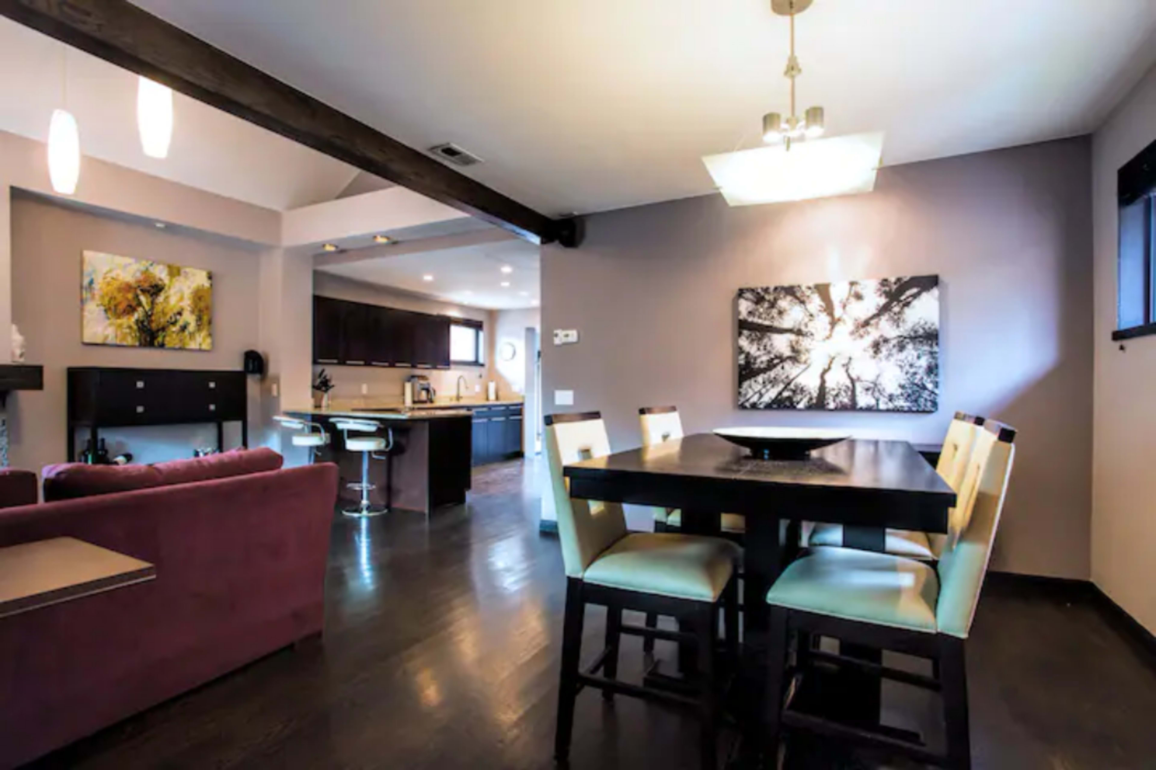 The image shows a modern dining area with a wooden table and high chairs adjacent to a living space featuring a purple couch and a kitchen in the background.