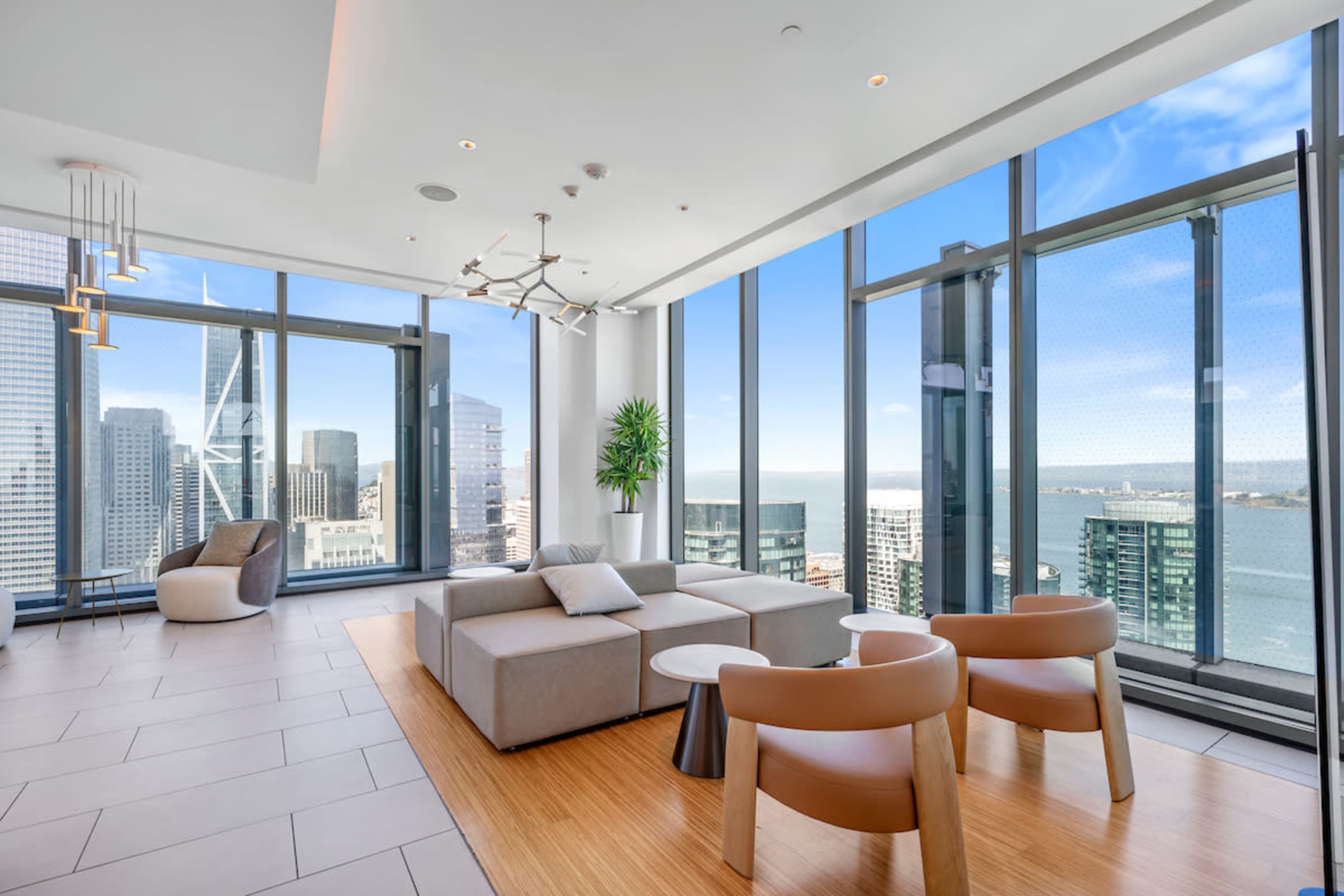 The image shows a modern living room with large windows overlooking a cityscape and the ocean, featuring a comfortable seating area and minimalist decor.