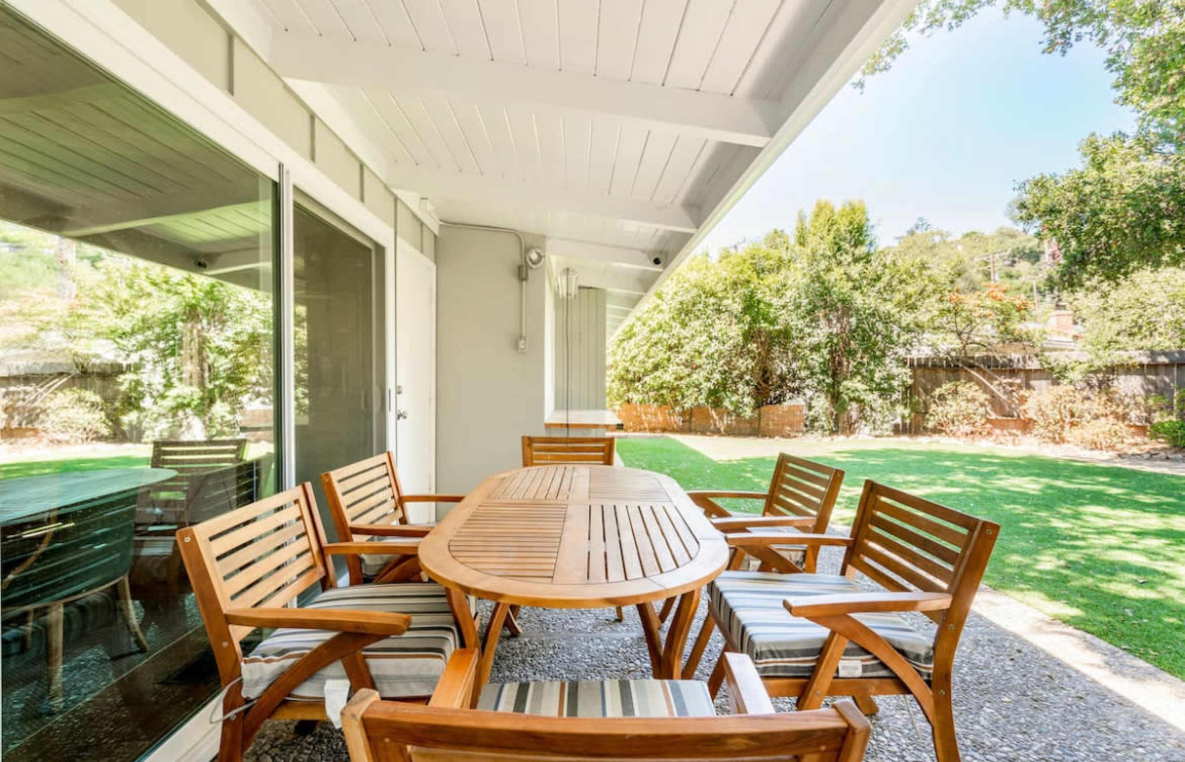 A wooden dining table surrounded by six chairs on a patio, overlooking a grassy yard and trees.