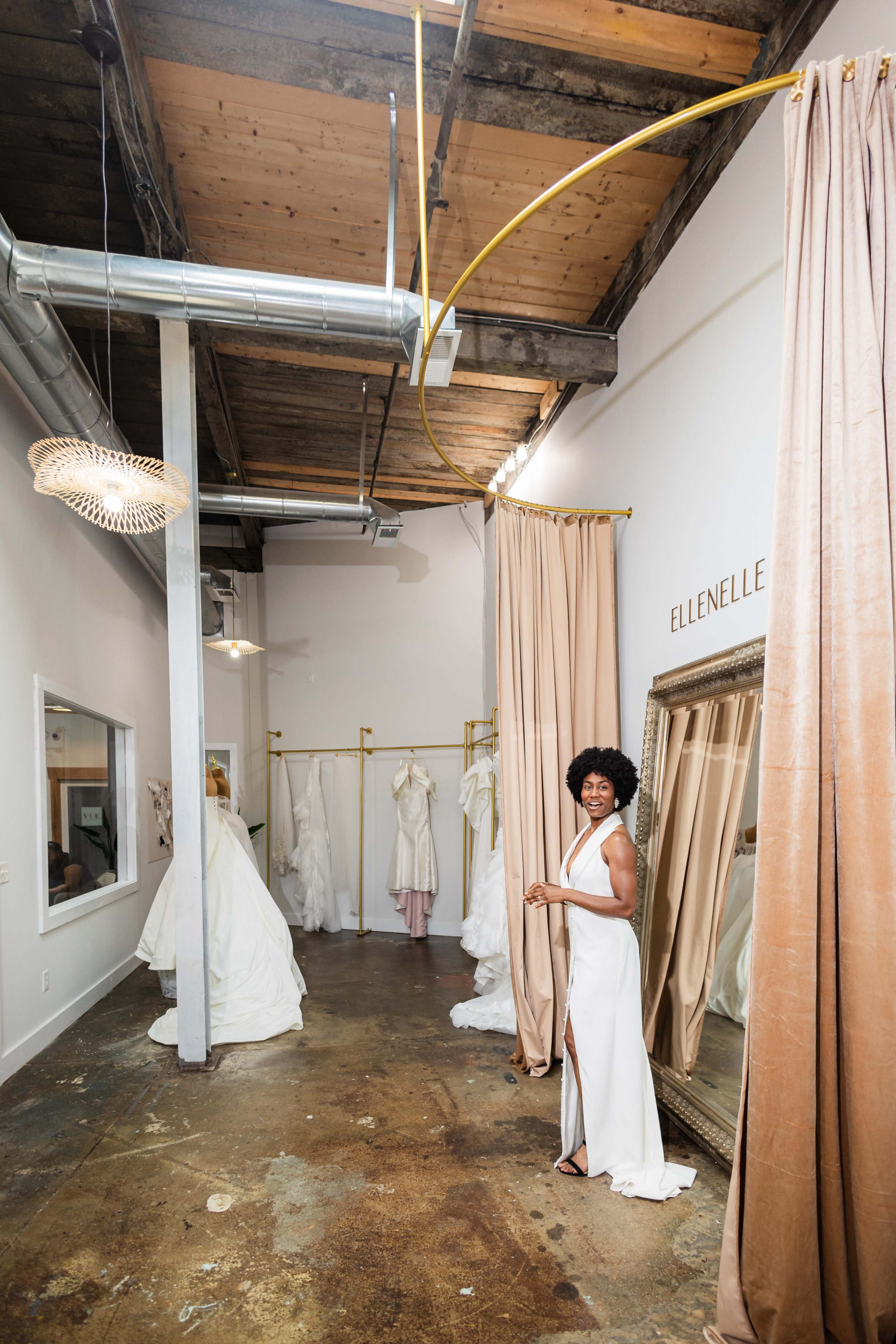 A woman in a white dress stands in a bridal salon featuring hanging garments and soft lighting.