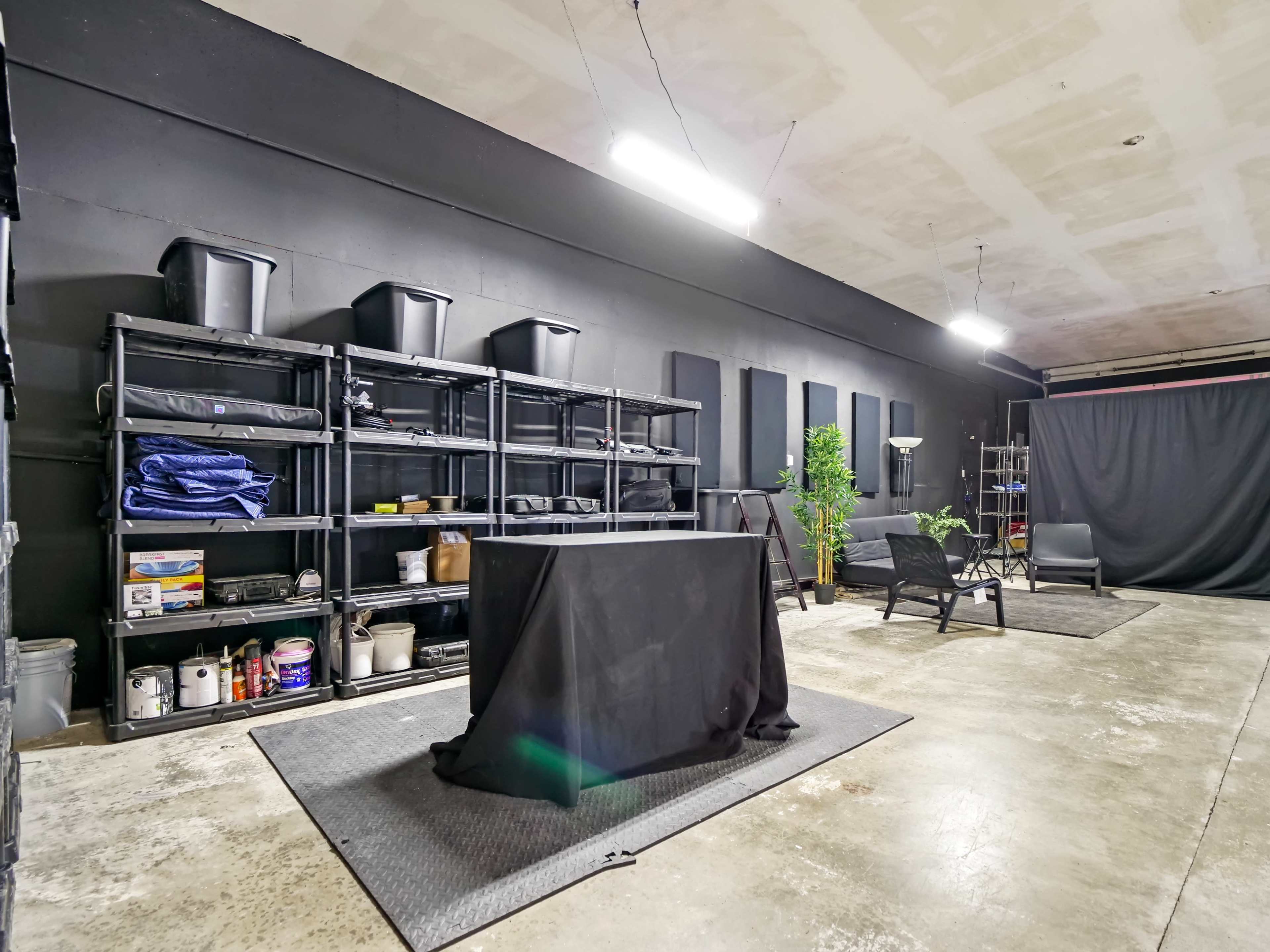 An industrial-style room with black walls, shelves filled with storage bins, a covered table in the center, and seating areas with minimal decor.
