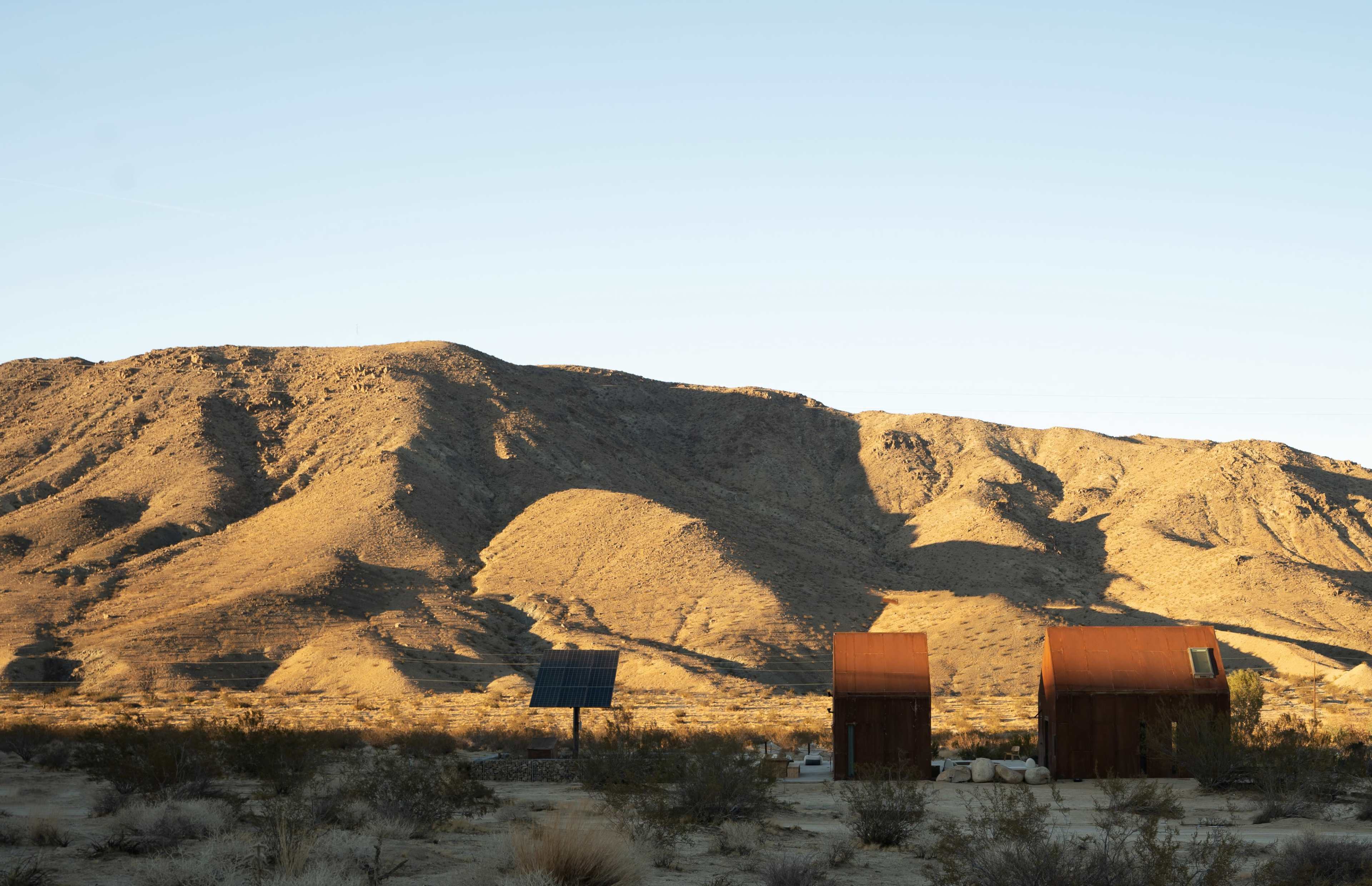 A pair of rusted metal structures are positioned in a desert landscape with a solar panel and distant mountains.