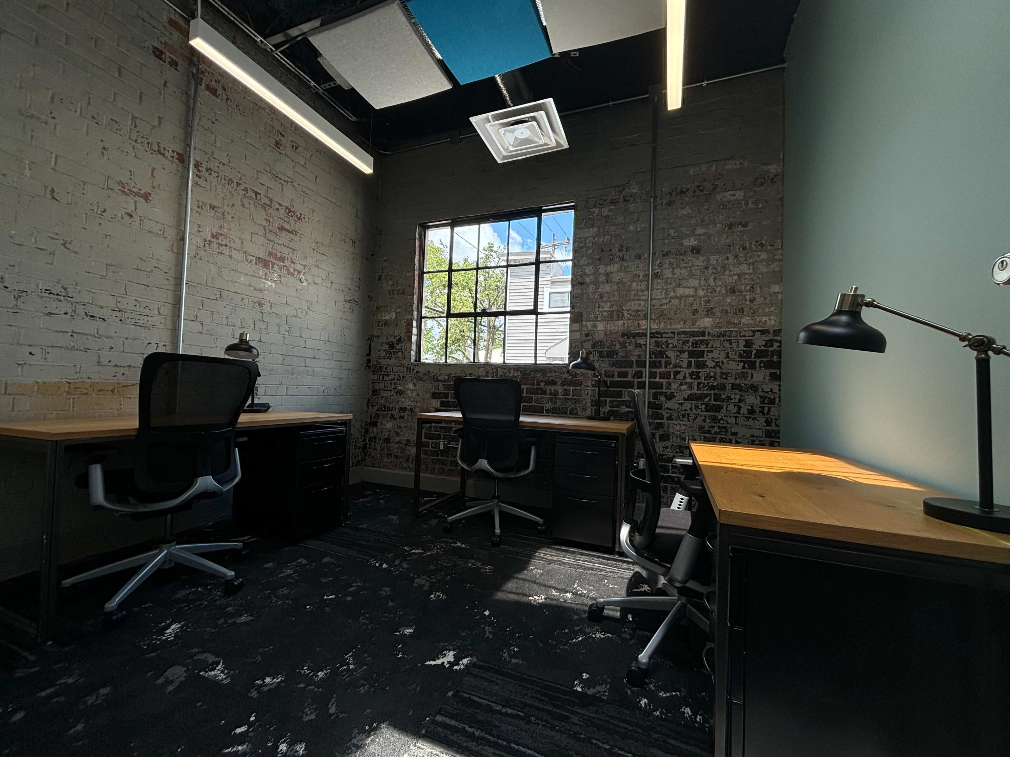 A well-lit office space with two desks and ergonomic chairs, featuring exposed brick walls and a large window allowing natural light to enter.