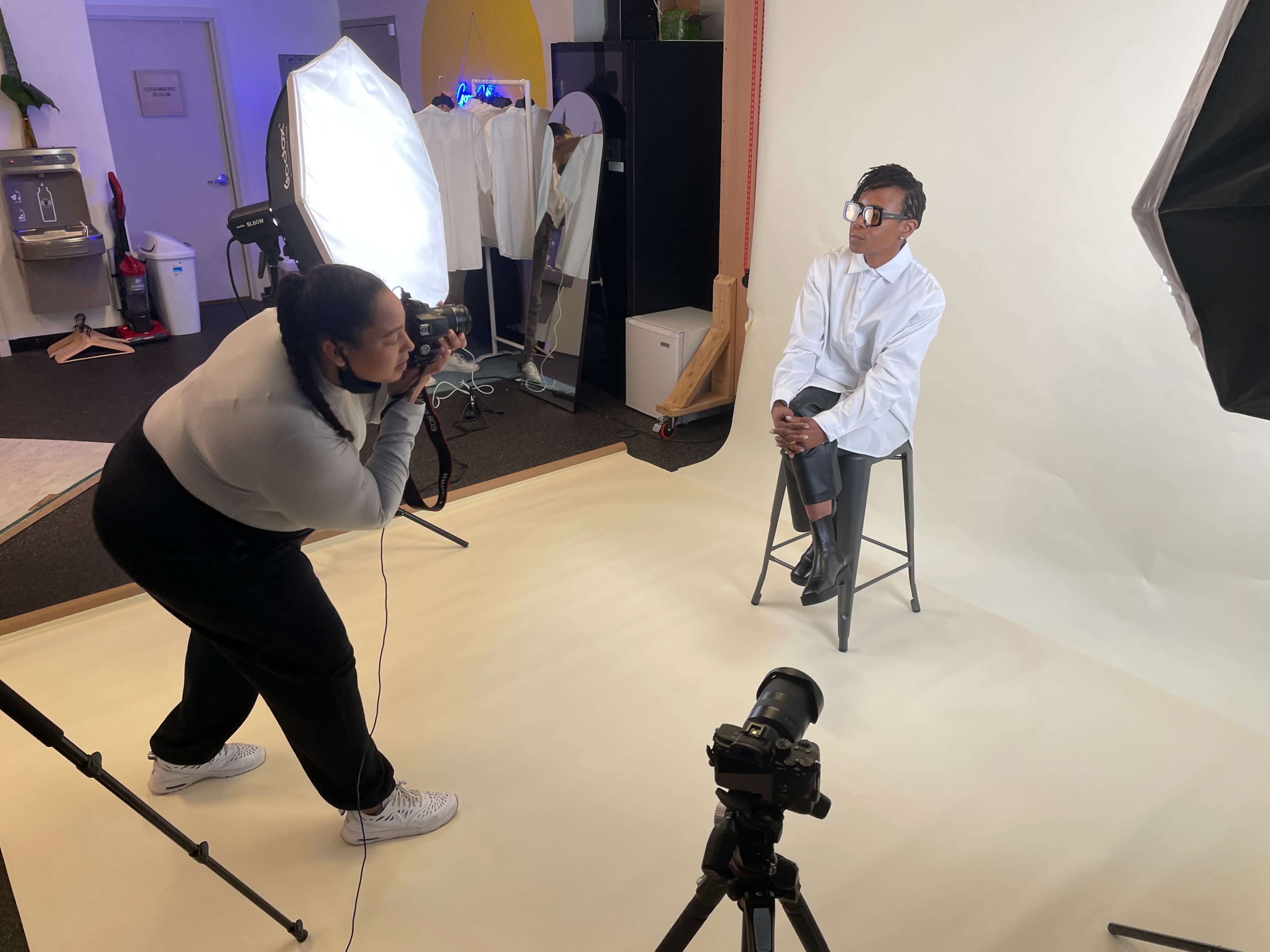 A photographer is taking a portrait of a model sitting on a stool in a studio with professional lighting equipment set up.