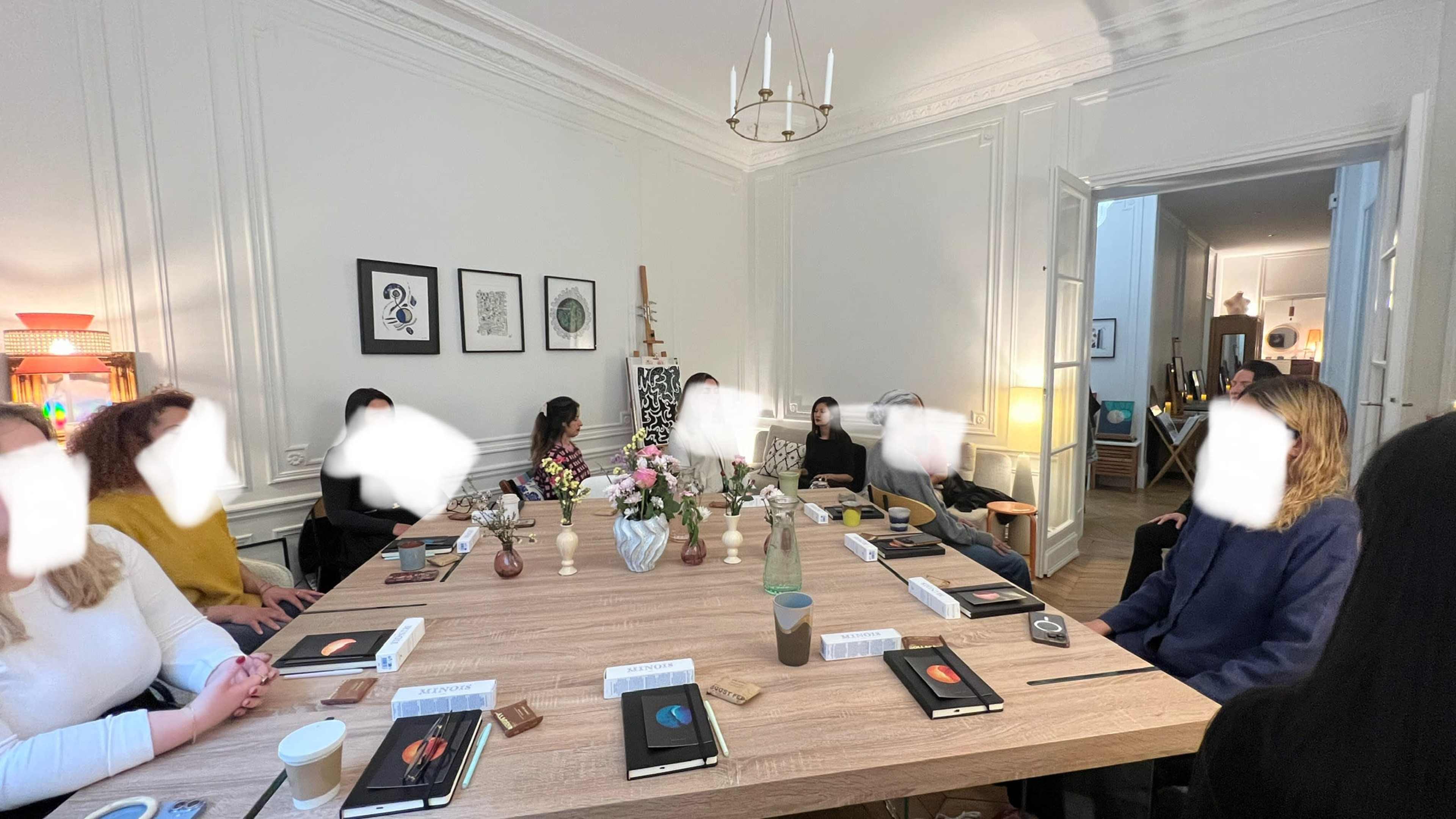 A group of people is seated around a large wooden table, engaged in conversation, with various drinks and snacks placed on the table amid decorative items.