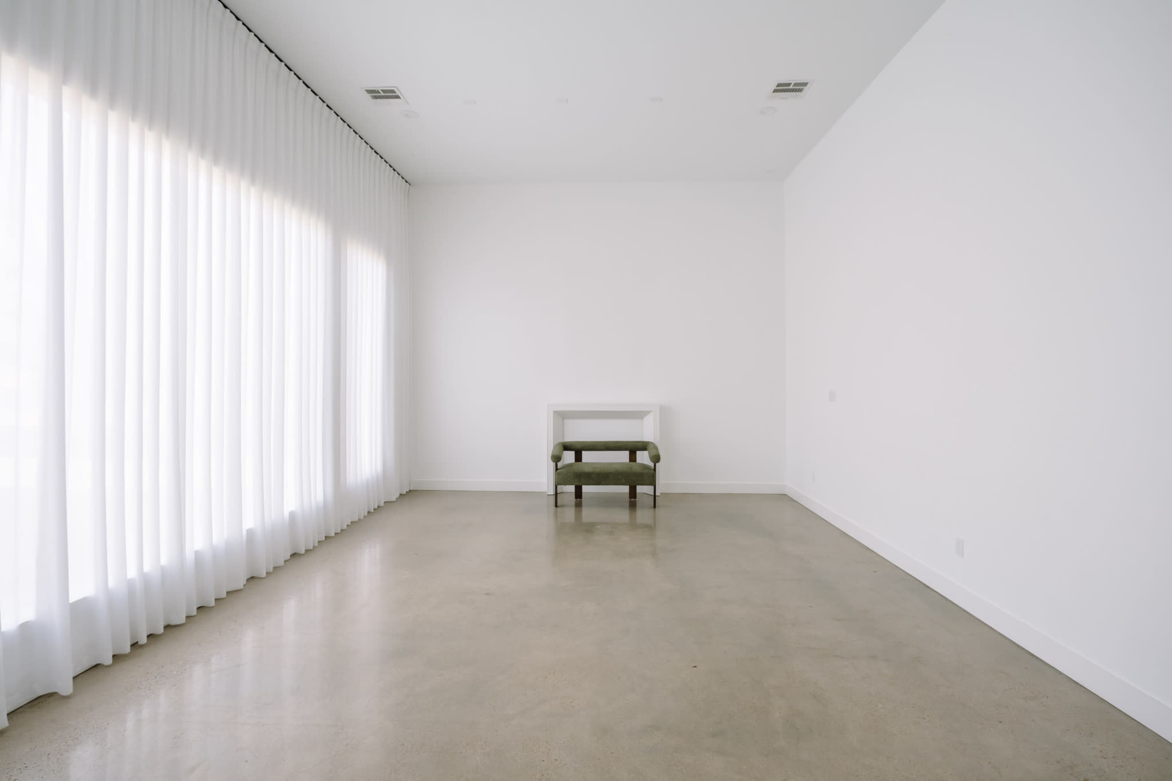The image shows a minimalist room with a polished concrete floor, a white wall, a small white table, and an olive green chair near large sheer curtains.