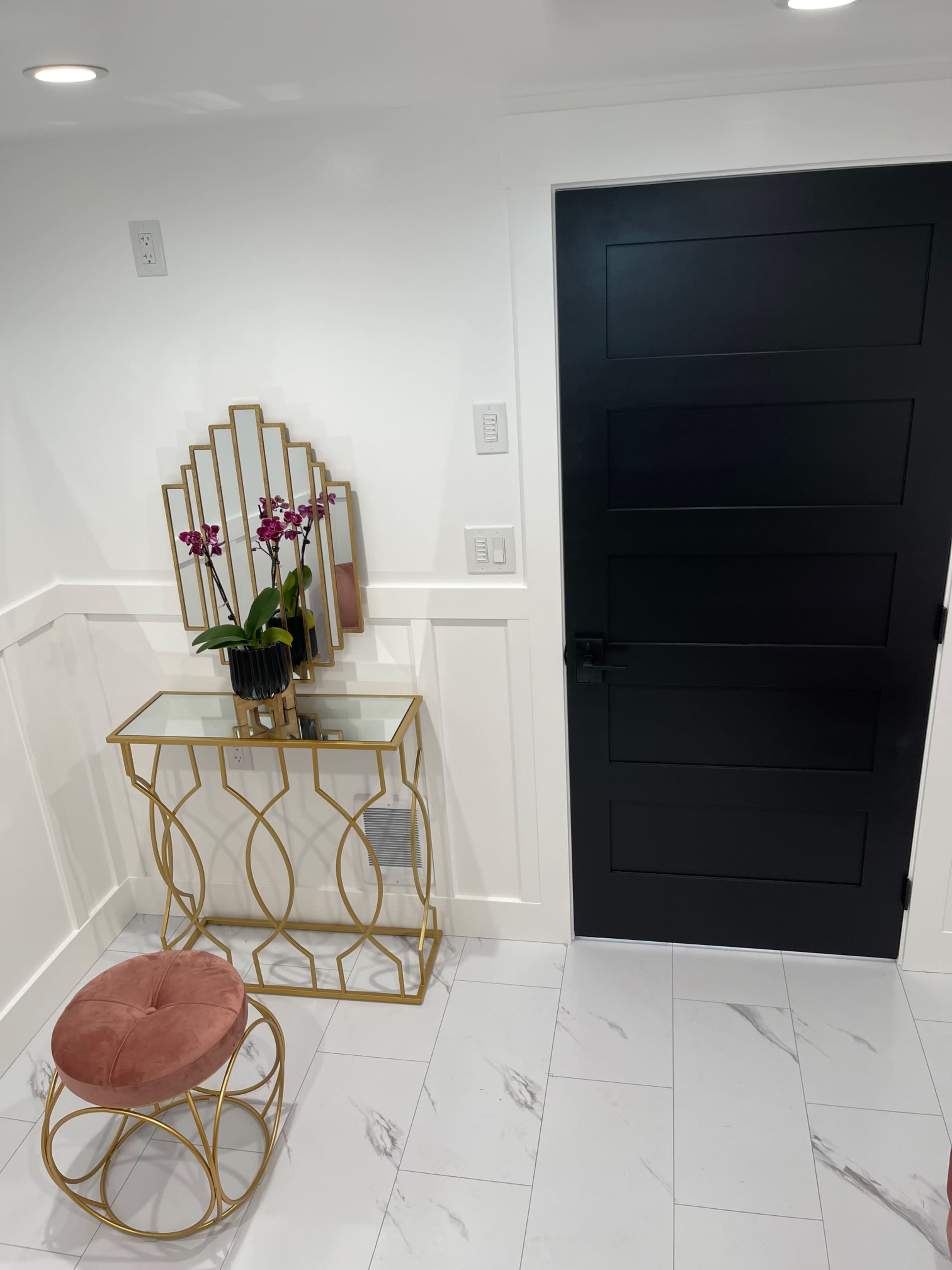 A modern entryway featuring a black door, a mirrored wall decoration, a gold accent table, a black potted plant with purple orchids, and a pink velvet stool.
