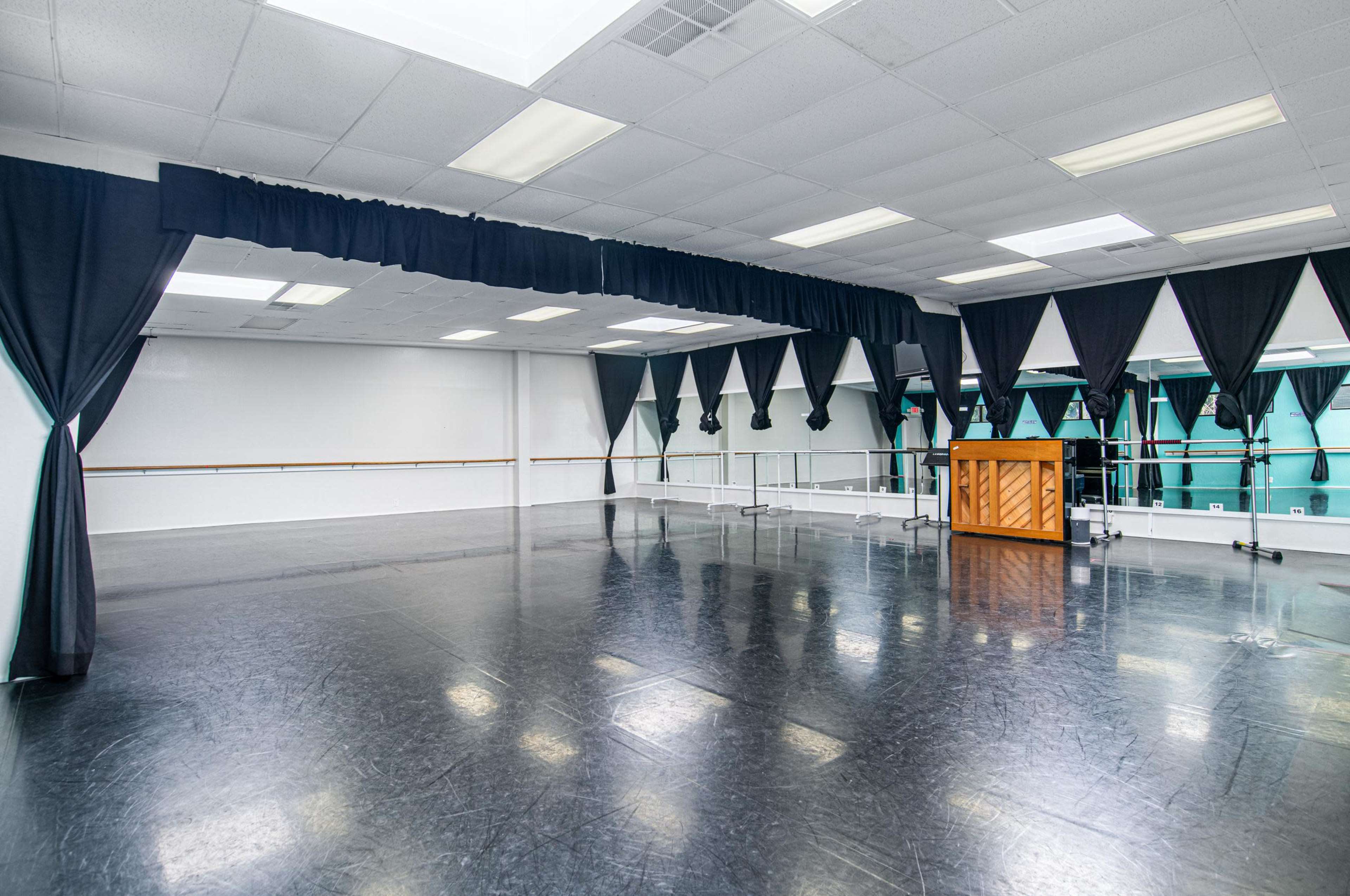 A spacious dance studio with dark flooring, large mirrors, and draped curtains, alongside a wooden piano positioned in the center.