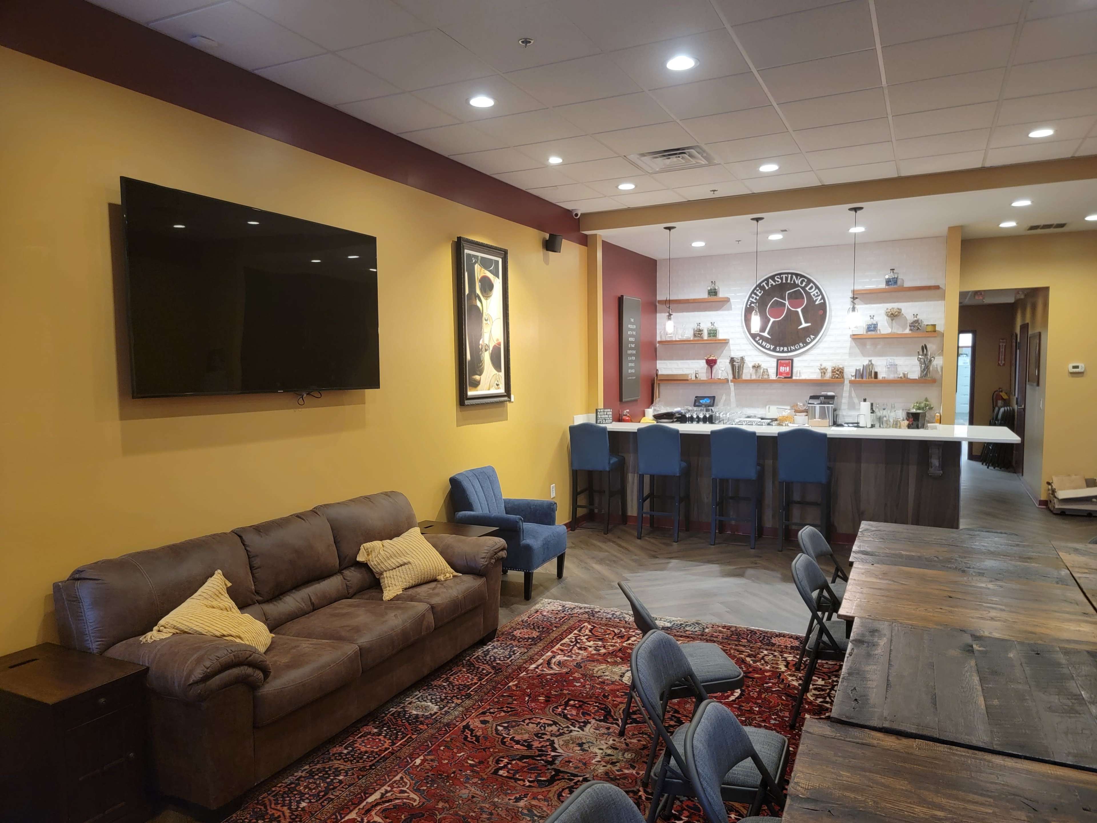 The image shows a modern, well-lit common area with a brown couch, blue chairs, a large television on the wall, and a kitchen in the background.