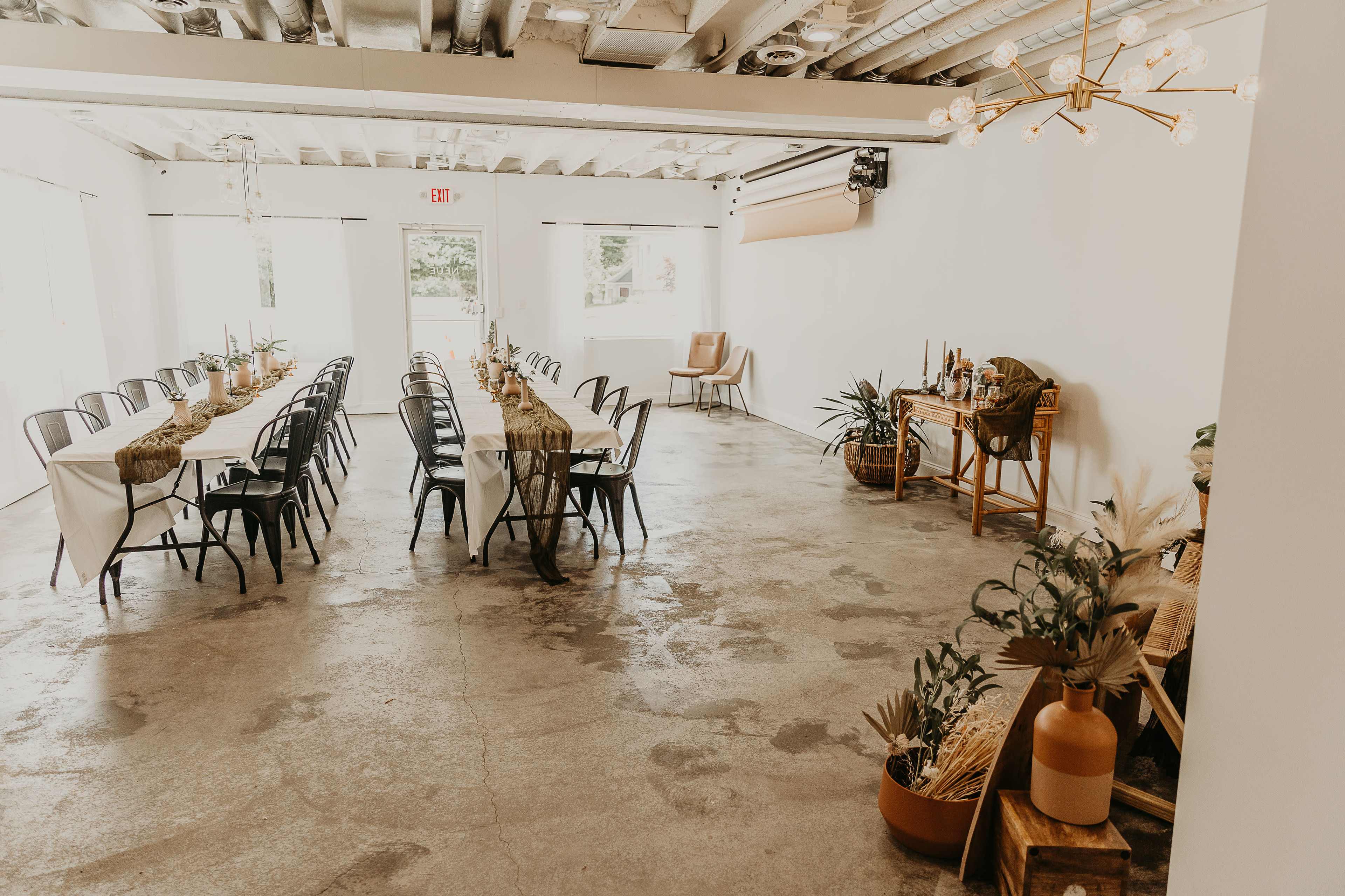 A spacious event room features long tables set with chairs, decorated with natural elements, and a refreshment area with plants.