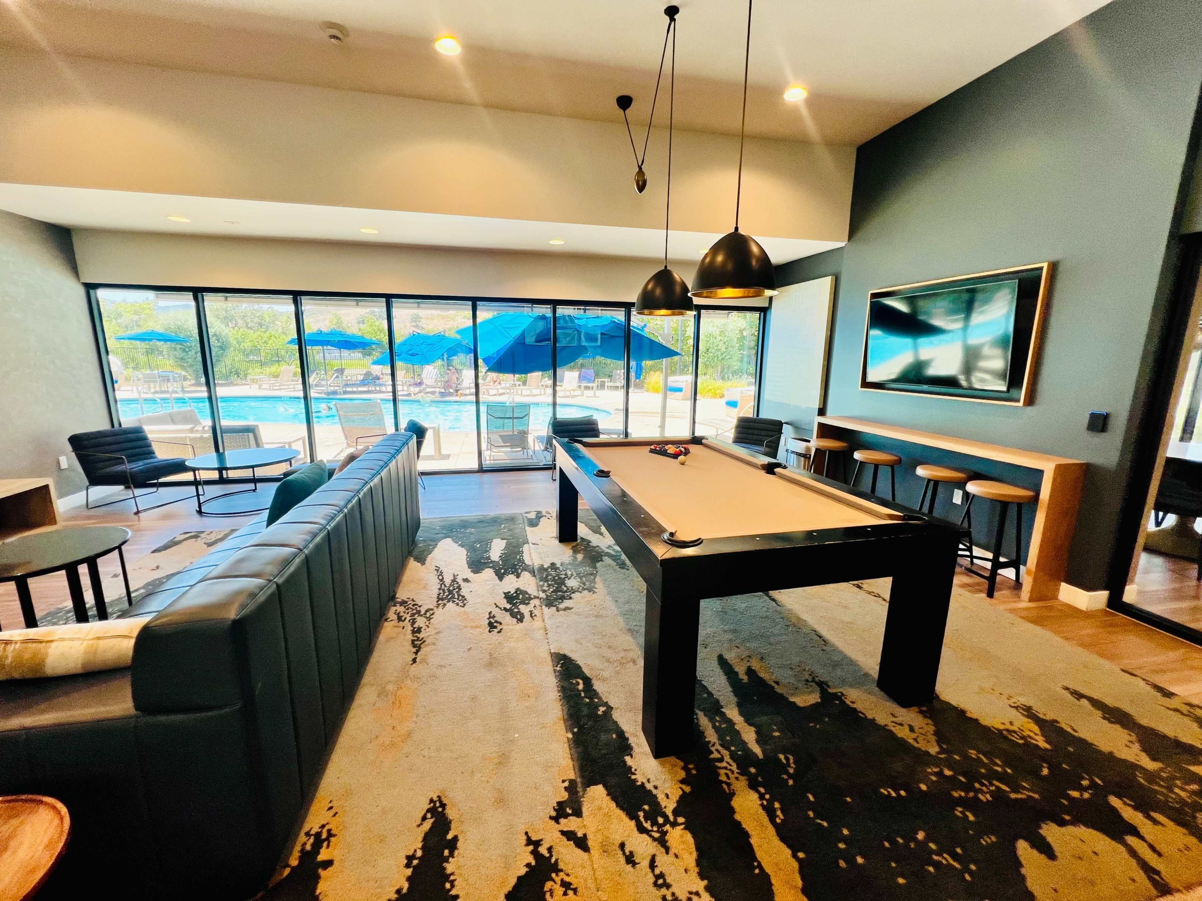 A lounge area with a pool table, seating, and large windows overlooking a pool and outdoor umbrellas.
