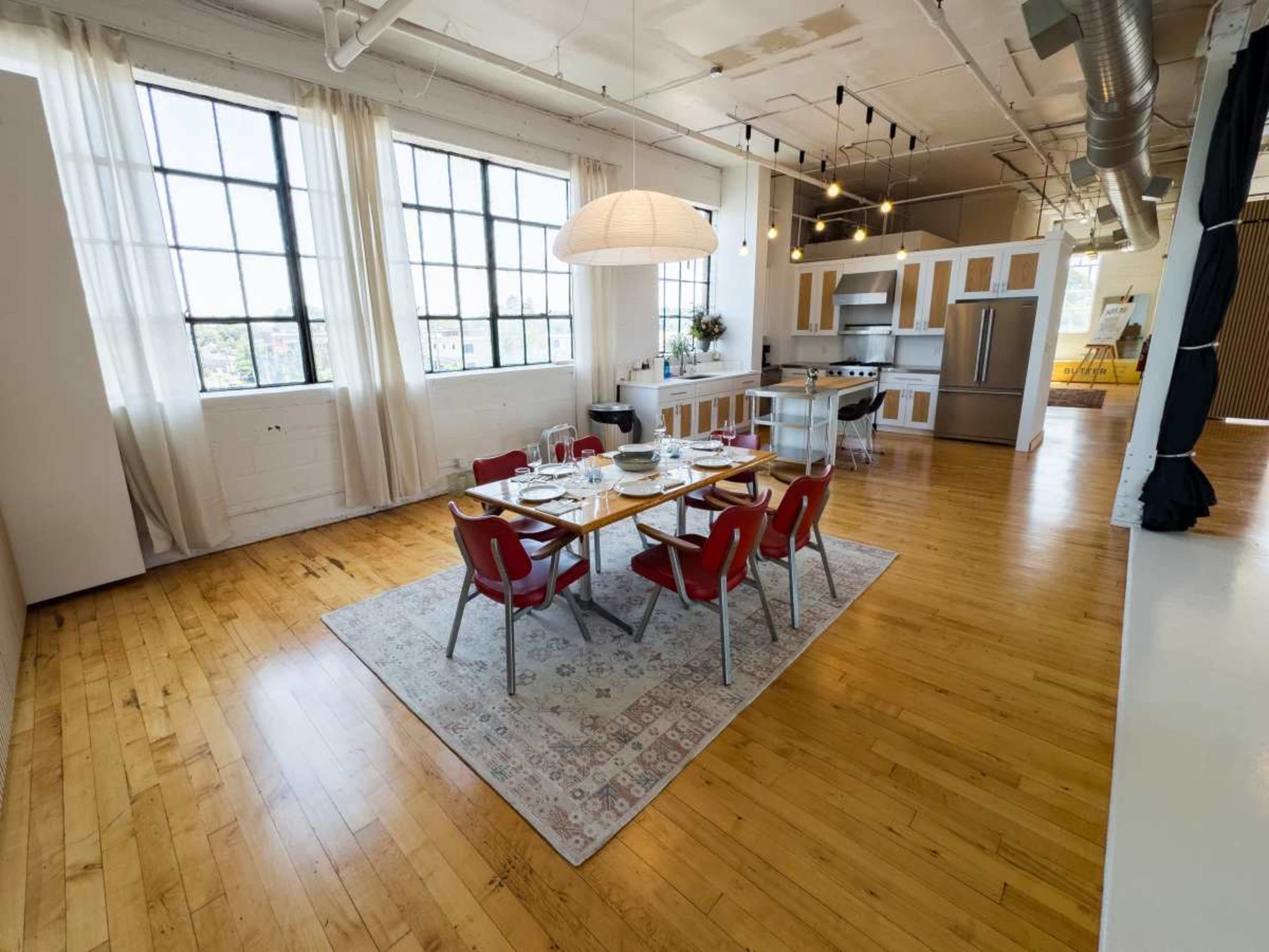 The image shows a bright and open dining area with a wooden table set for a meal, surrounded by red chairs, and a kitchen visible in the background.