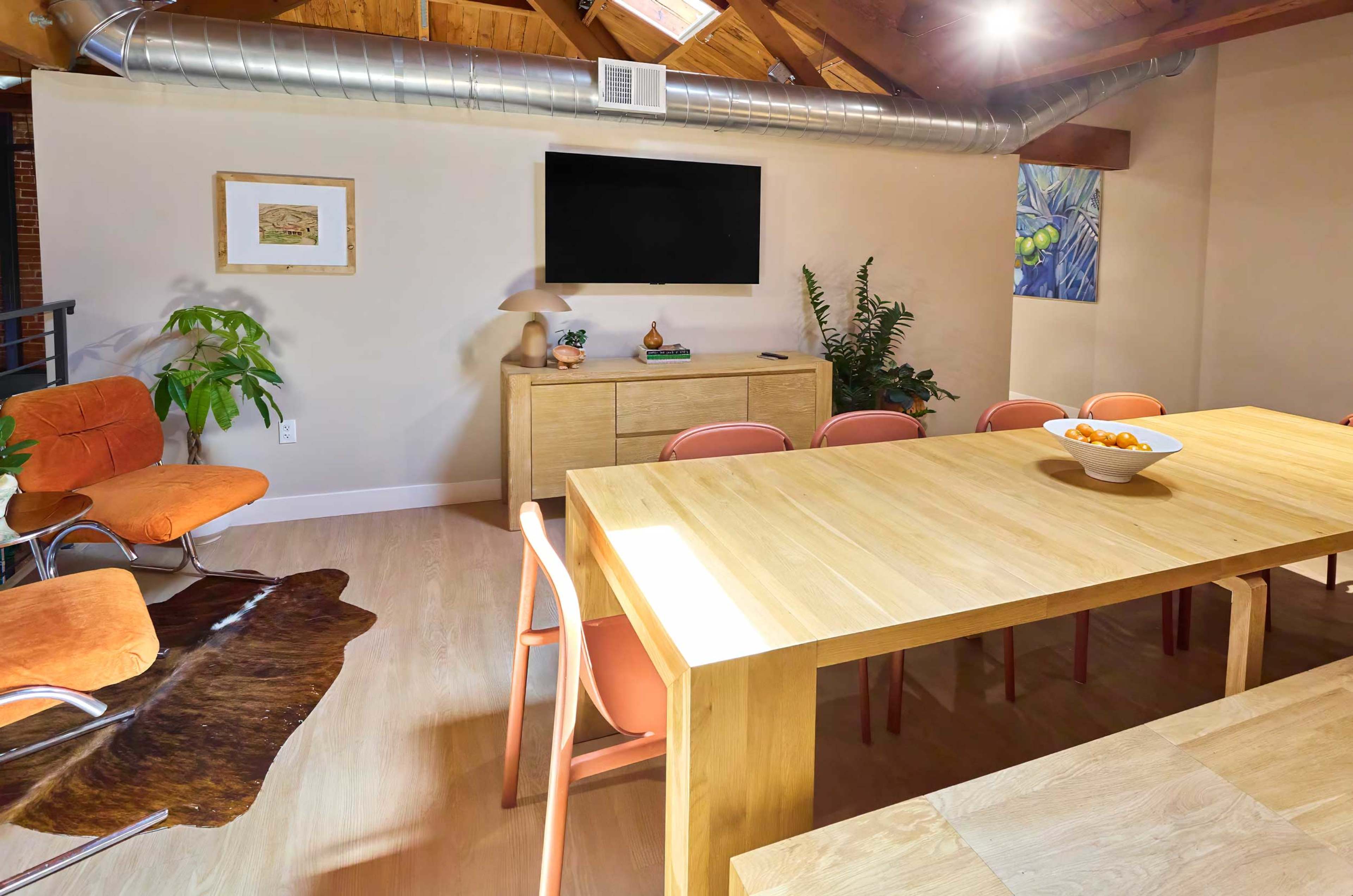 The image shows a spacious dining area with a wooden table, pink chairs, a sideboard, a television mounted on the wall, and various indoor plants.