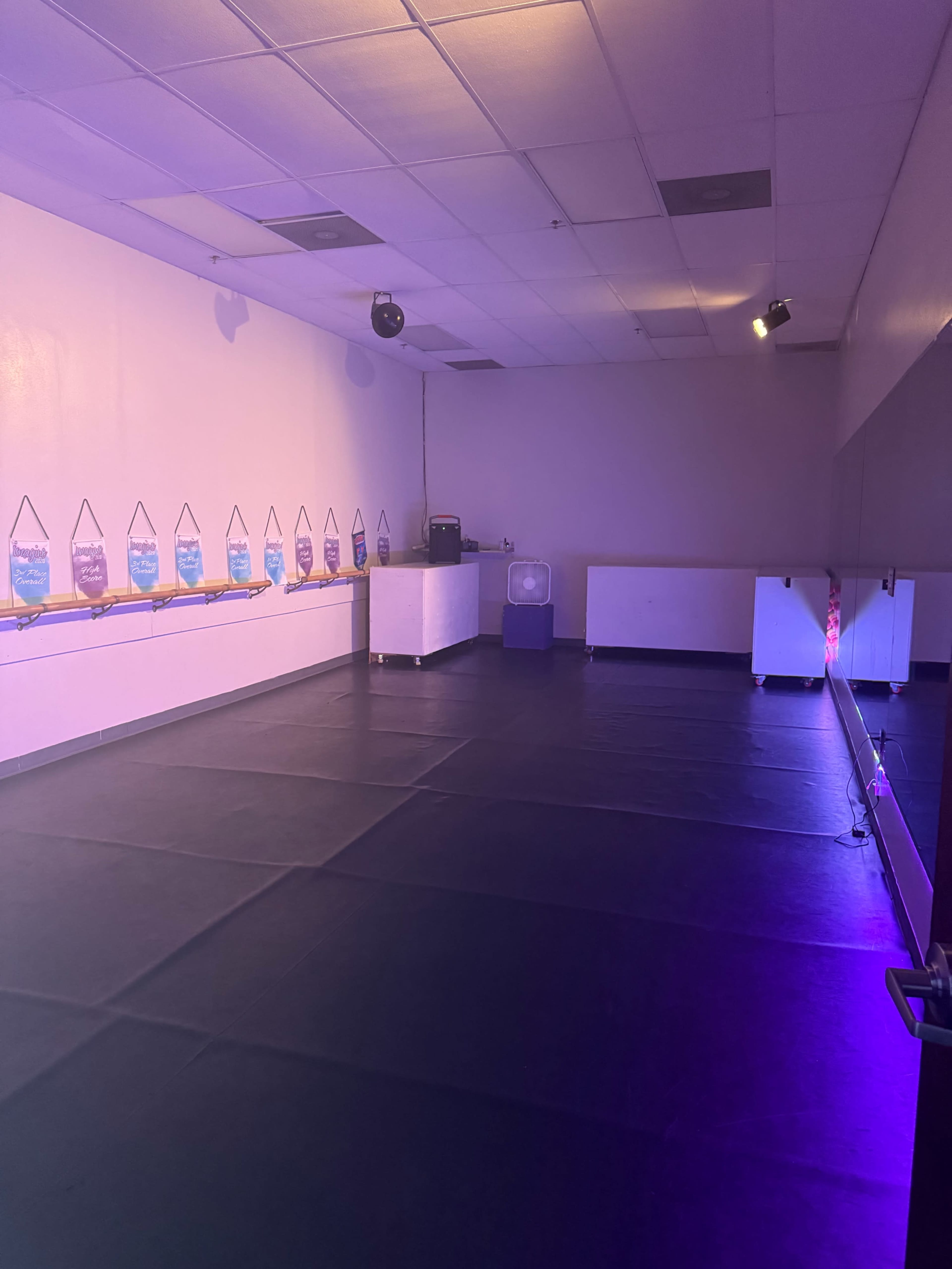 The image shows an empty room with black flooring, a row of translucent containers on a shelf, and a small fan in the corner.