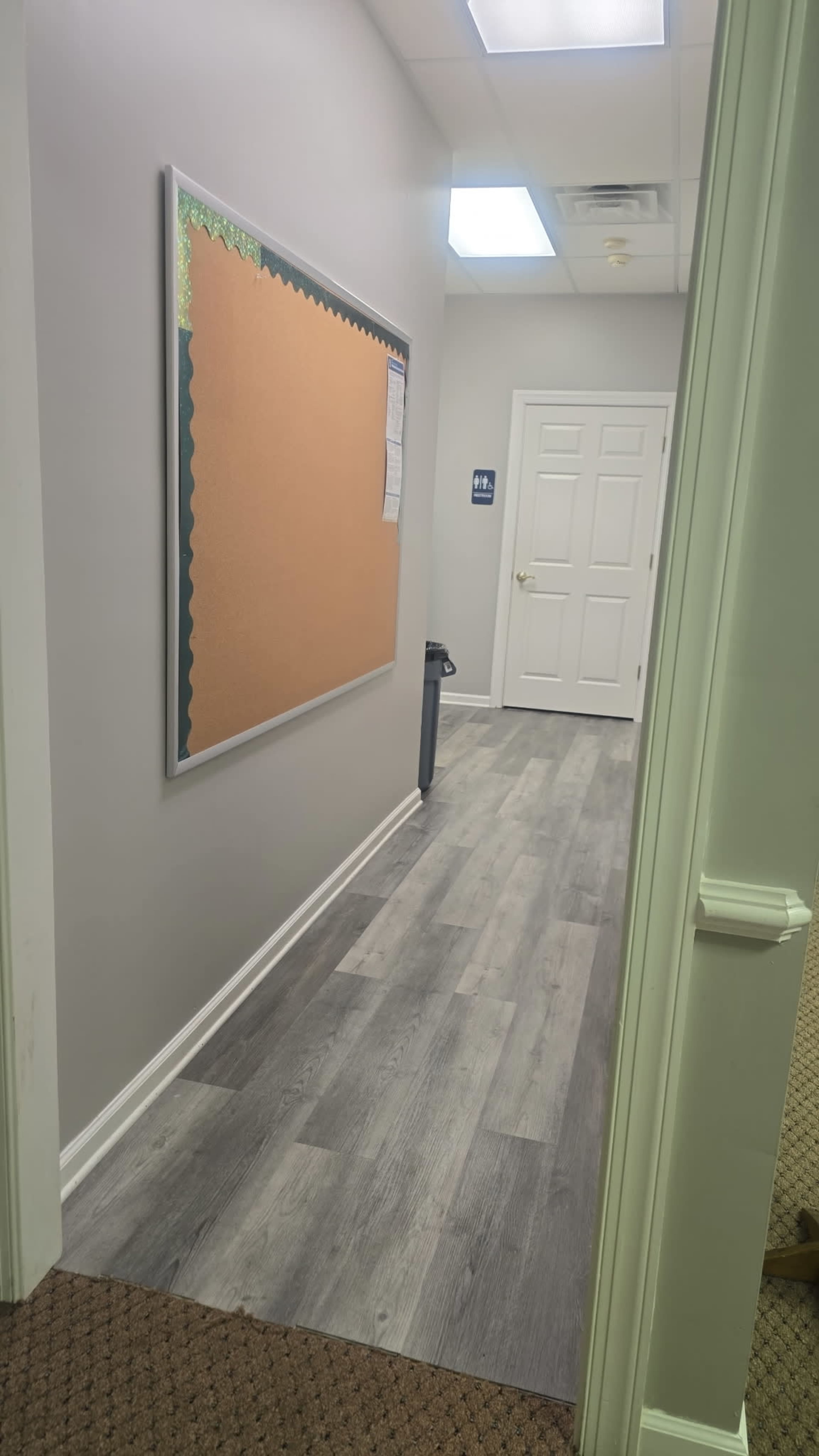 The image shows a hallway with a bulletin board on the left, a closed door at the end, and gray flooring that transitions to carpet at the entrance.