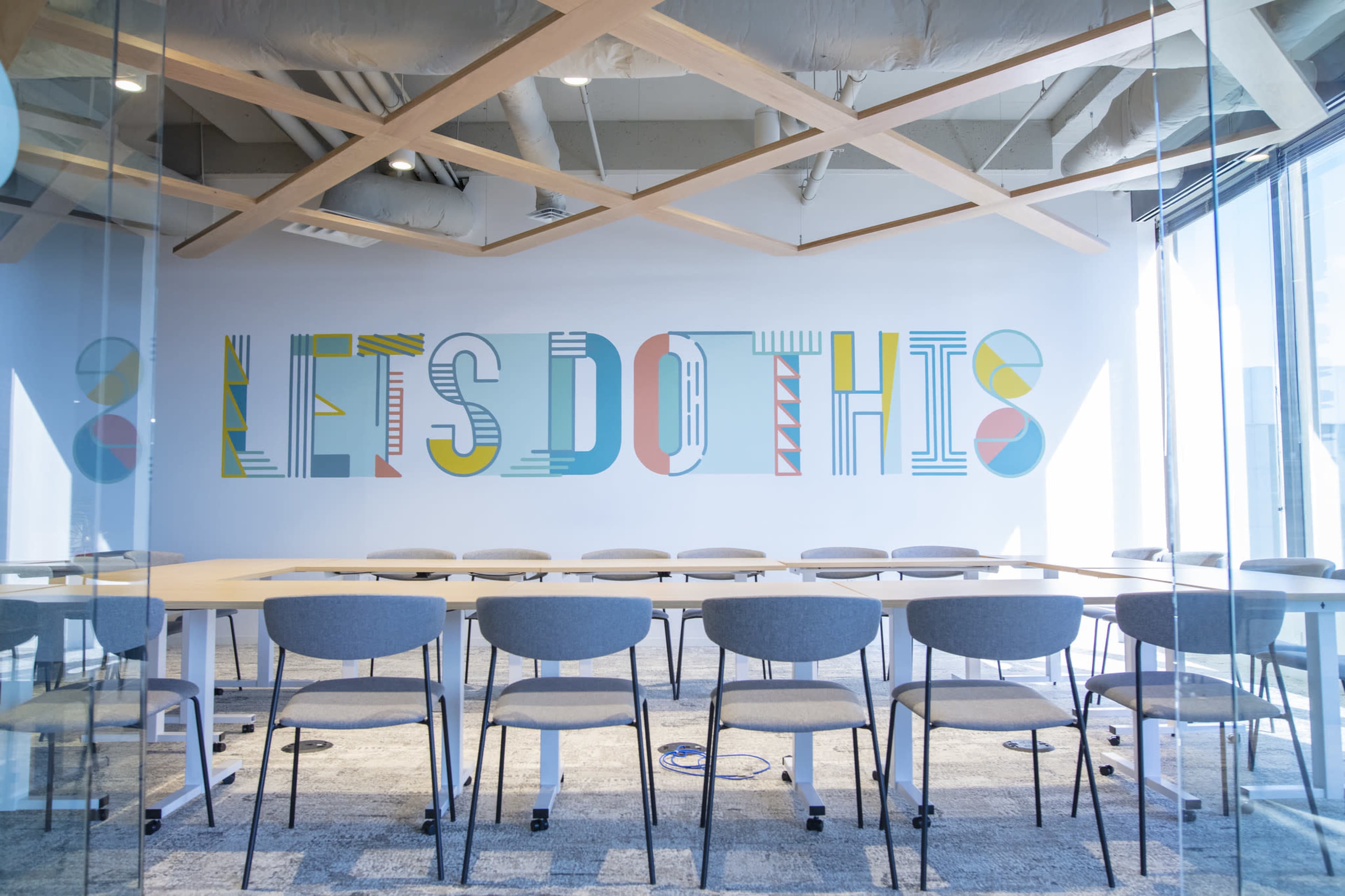 A modern conference room features a large decorative wall that says "LET'S DO THIS" in colorful letters, with a long table and several chairs arranged in the room.
