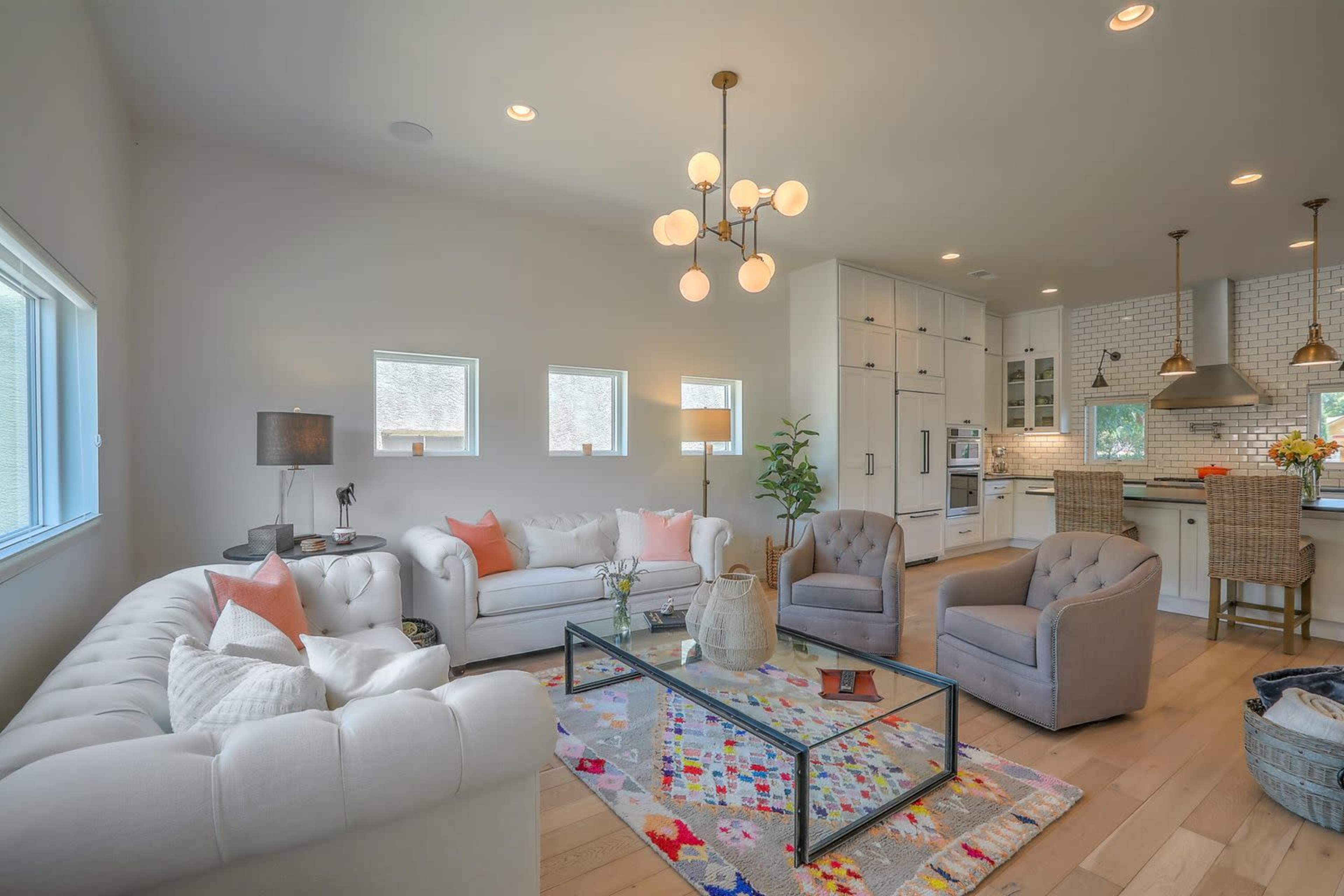 A modern living room features white sofas, a glass coffee table, and two gray armchairs, with a light fixture above and an open kitchen in the background.