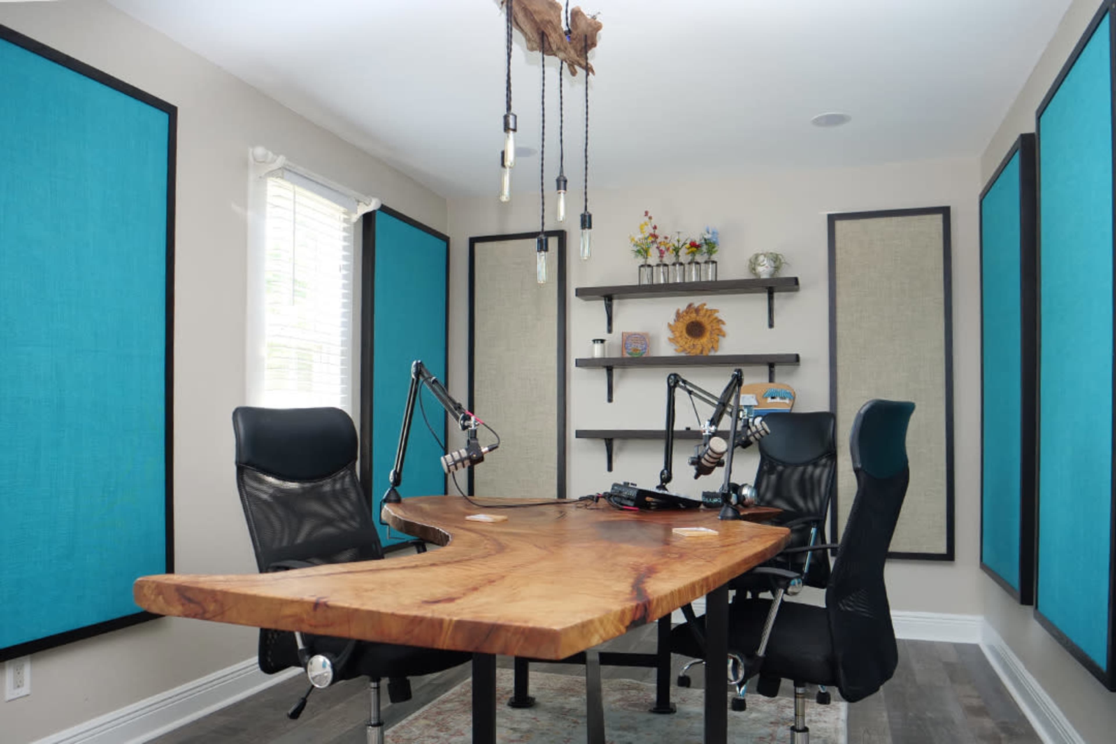 The image shows a modern podcast recording studio with a large wooden table, two black office chairs, and soundproof blue panels on the walls.