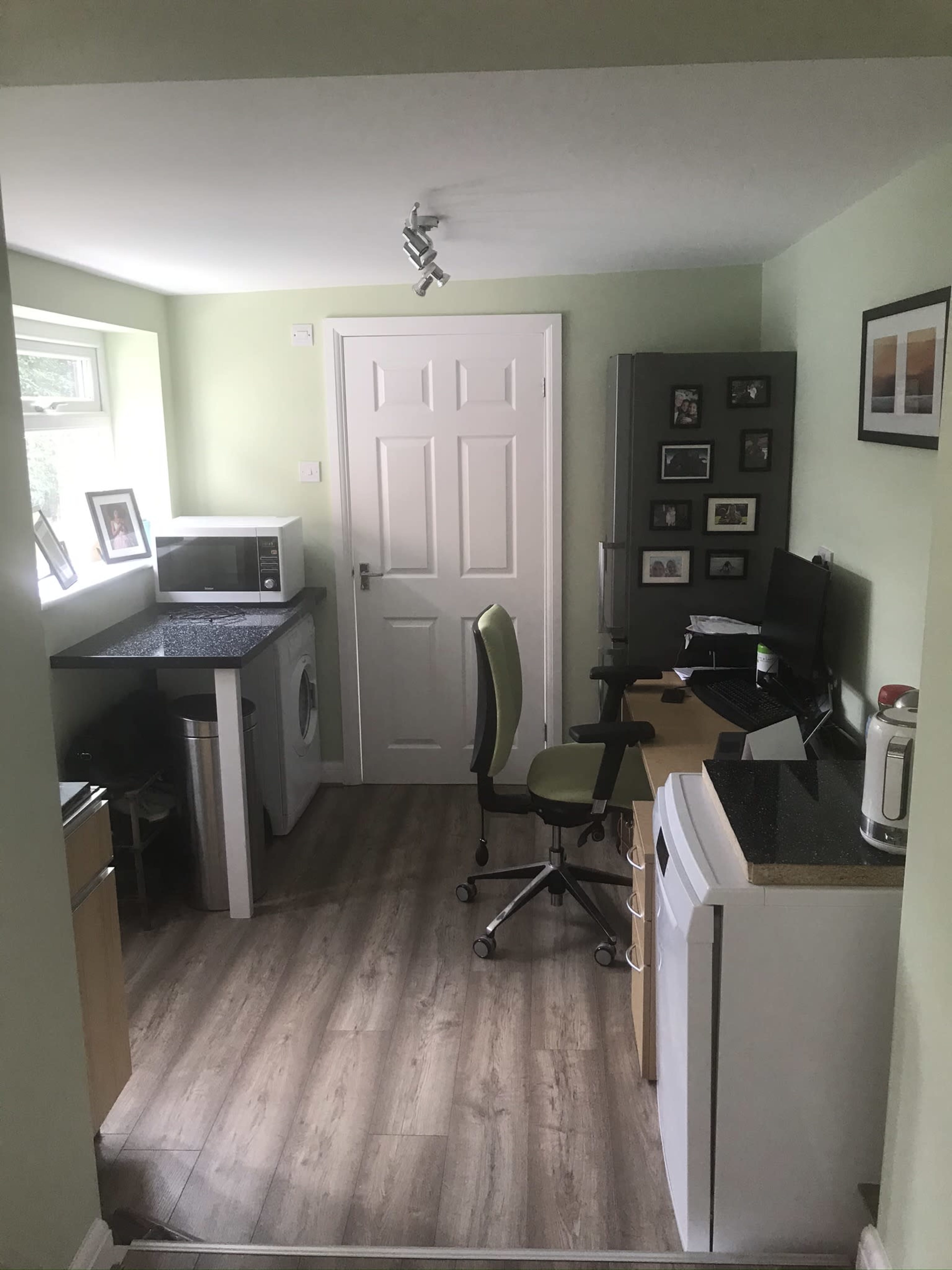 A small, functional kitchen area with a washing machine, microwave, refrigerator, and a desk with a computer and office chair.