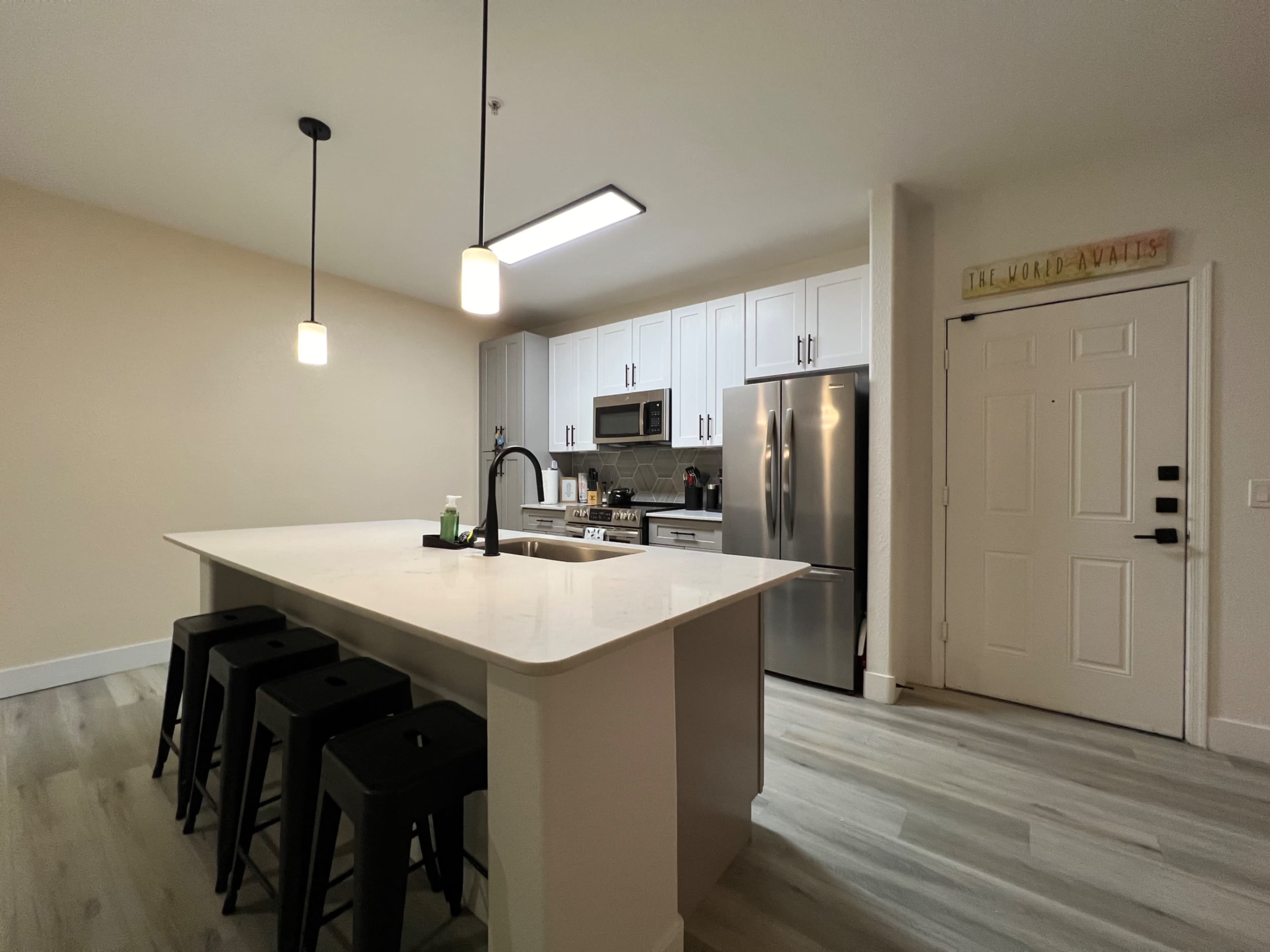 A modern kitchen with white cabinetry, a large island with seating, stainless steel appliances, and a front door visible on the right.