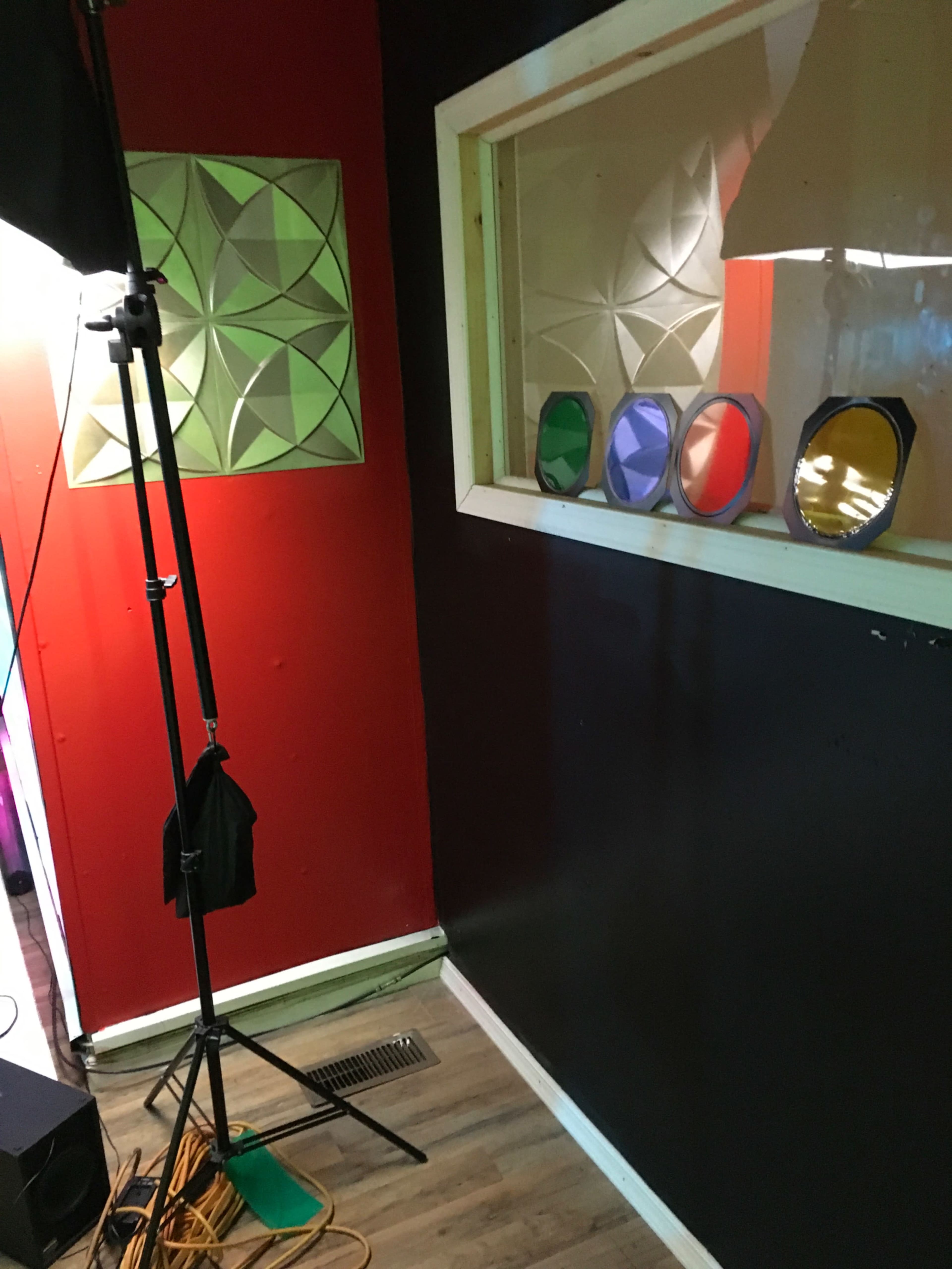 A corner of a room with a red wall, a green geometric panel, a window displaying colored glass panels, and a lighting stand.