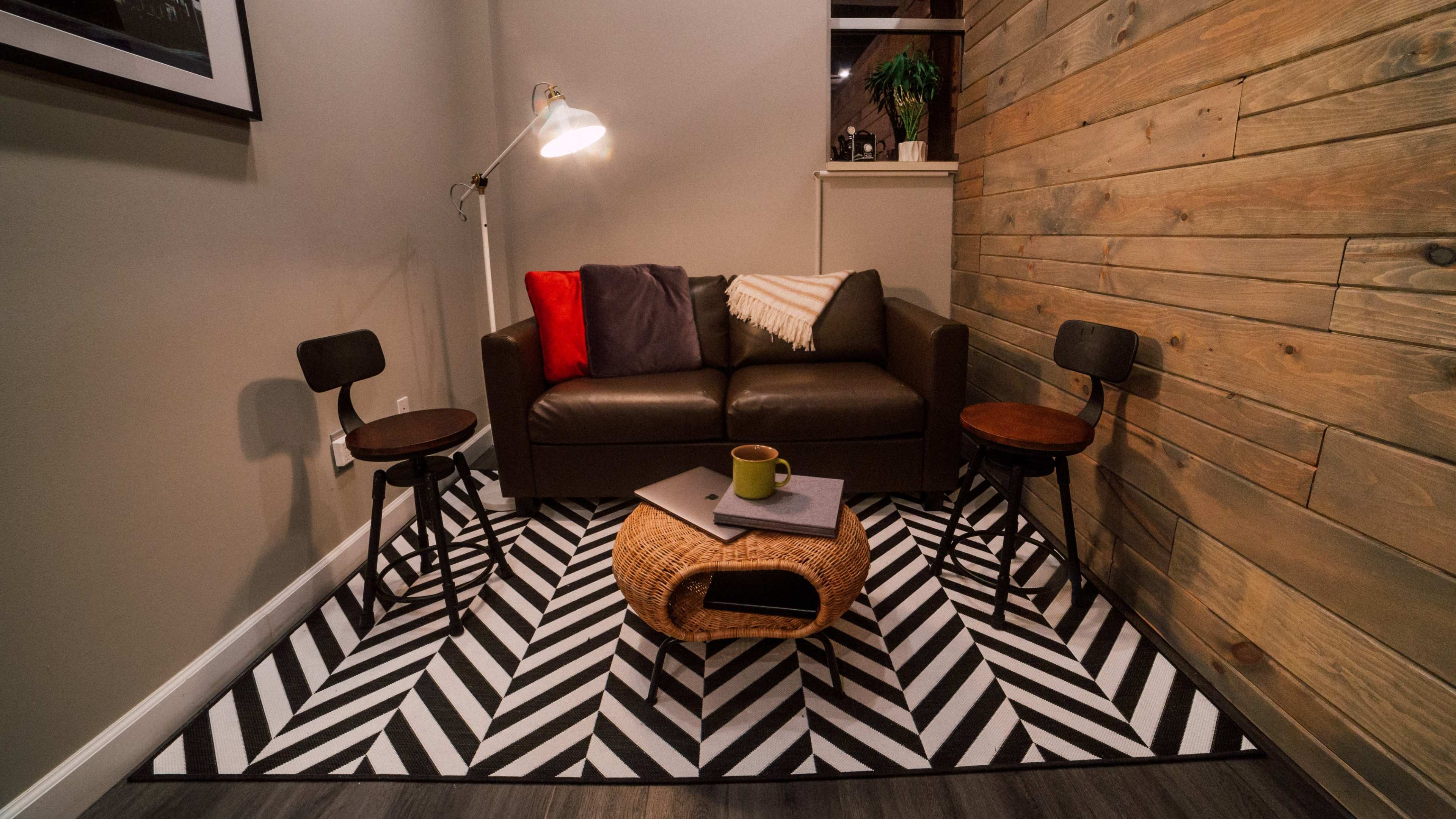 A cozy seating area featuring a brown sofa with cushions, two wooden side chairs, a round coffee table, and a patterned rug.