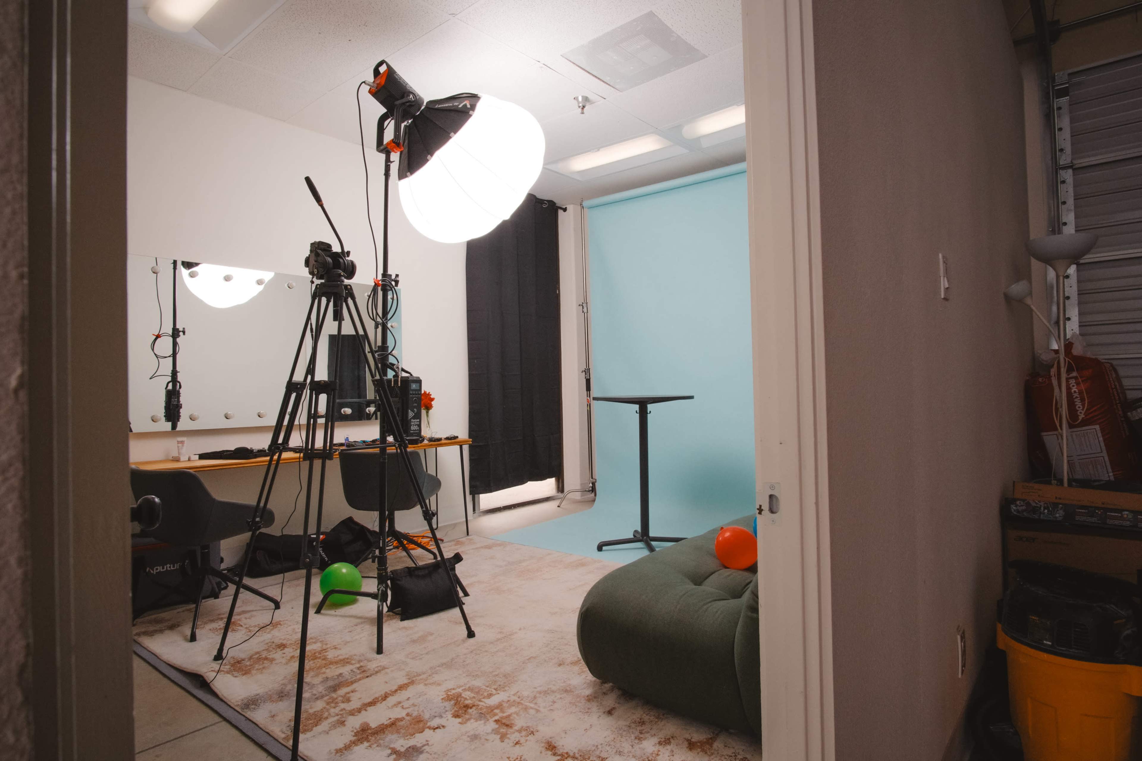 The image shows a small studio room with a light setup, camera on a tripod, a mirror with lights, a green chair, and a blue backdrop.